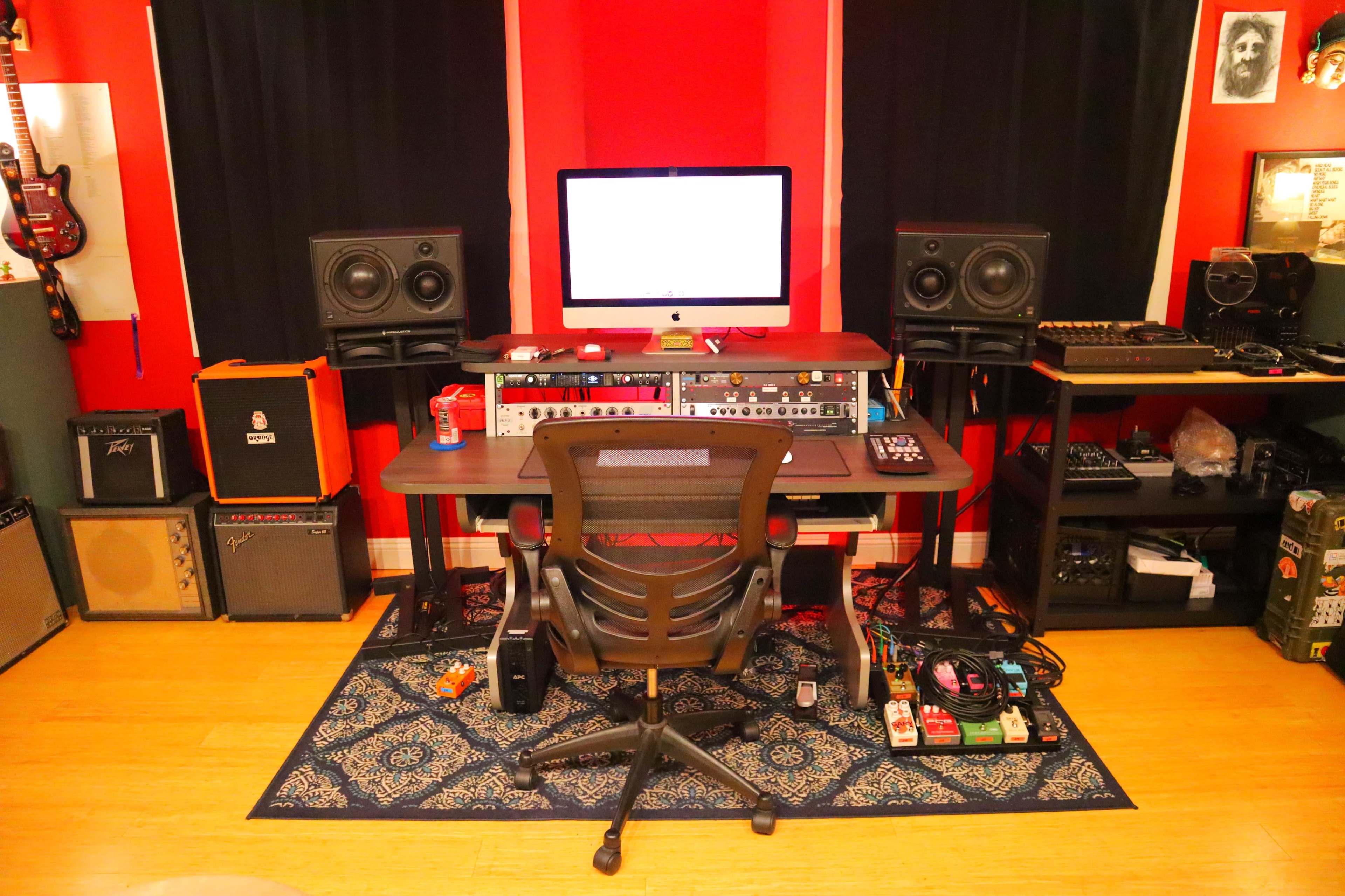 The image shows a music recording studio featuring a central desk with an iMac, audio interface, studio monitors, and various musical instruments and equipment surrounding it.