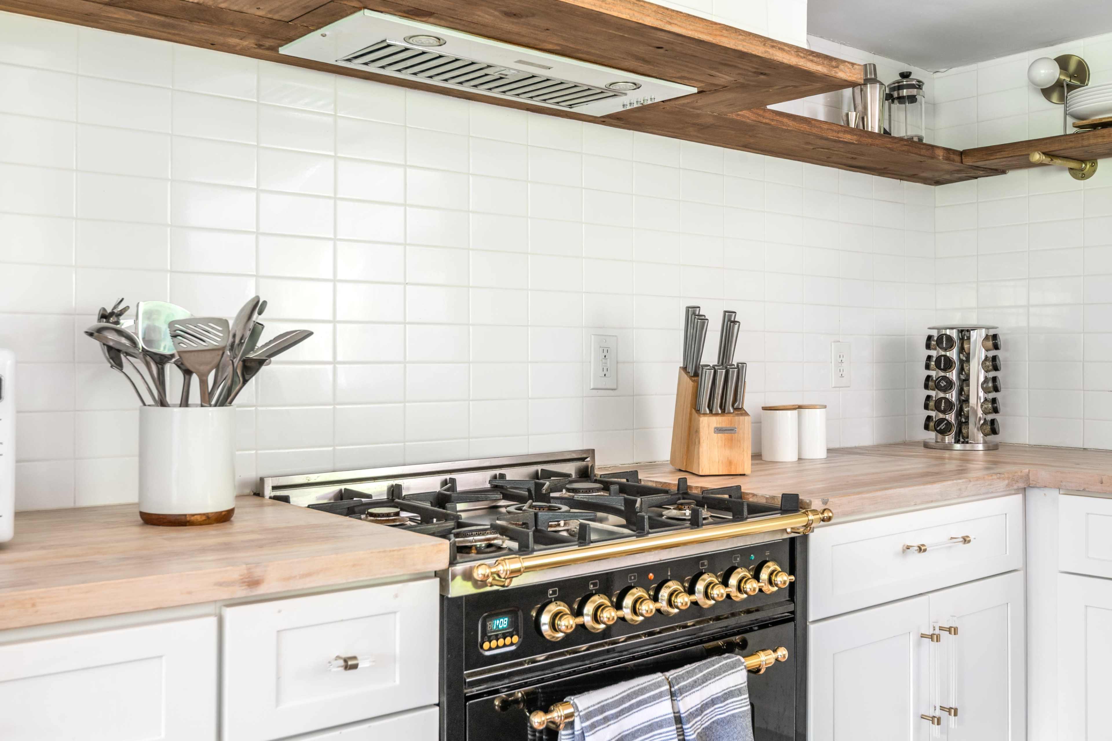 The image shows a modern kitchen with a black gas stove, white cabinets, wooden shelves, and various kitchen utensils displayed.