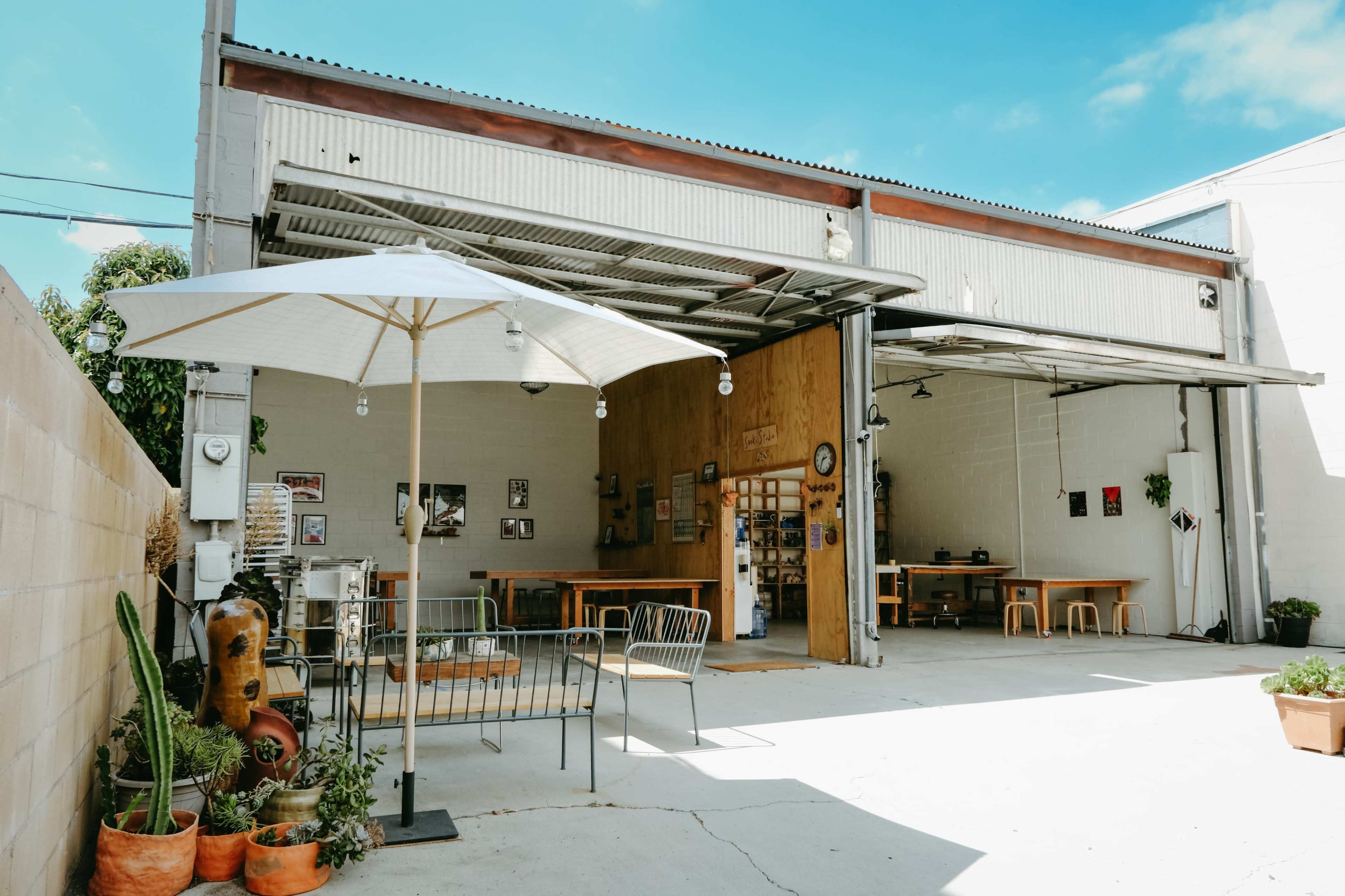 A spacious outdoor area of a café or workshop with a large umbrella, seating, and a few tables under a well-lit, open garage-style structure.