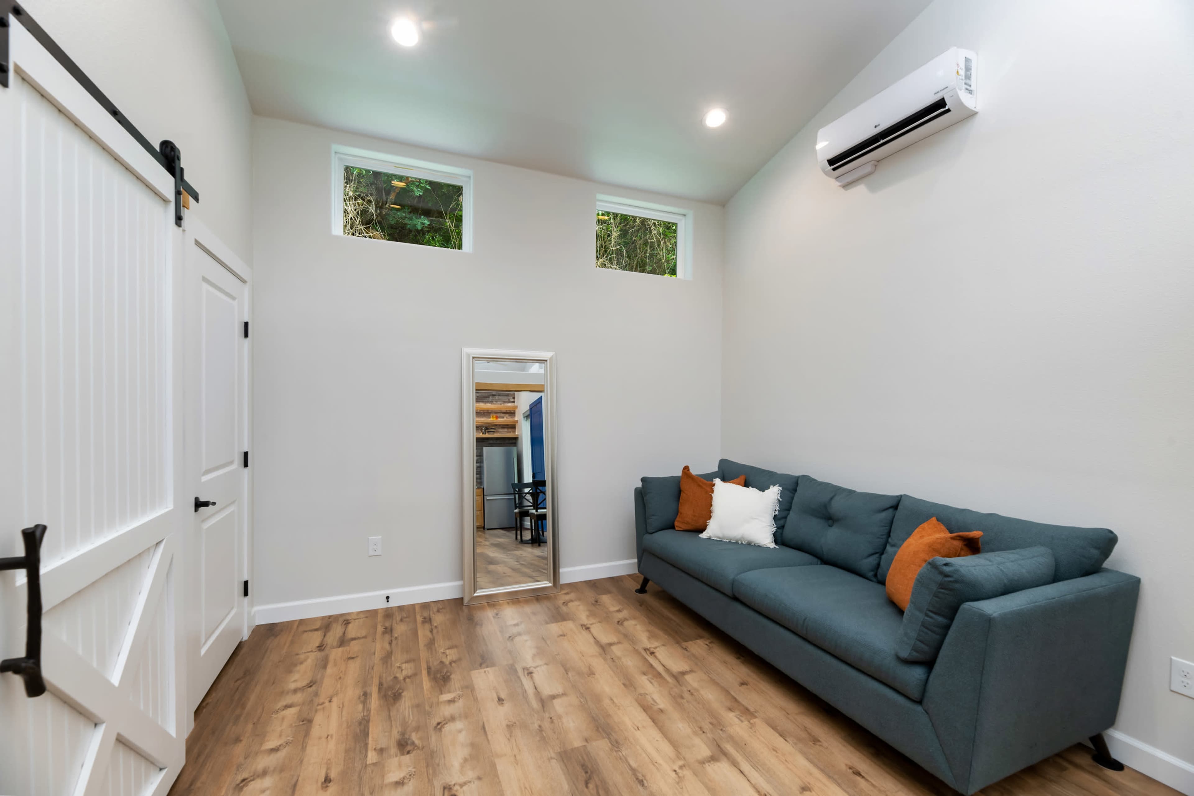 The image shows a small, modern living space with a couch, a large mirror, and windows allowing natural light to enter.