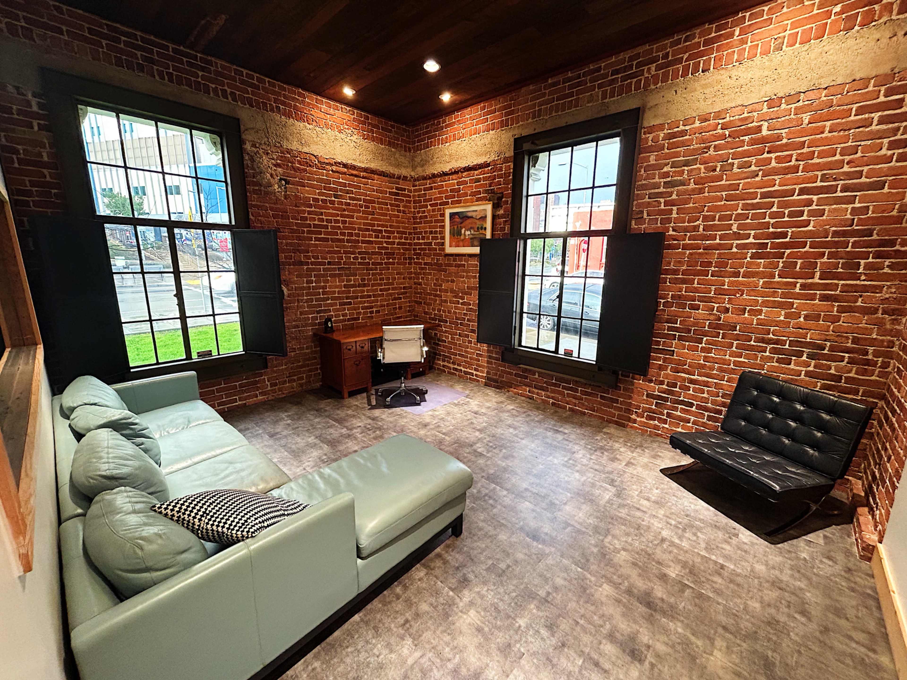 A small room with exposed brick walls, a light green sofa, a black chair, and a wooden desk near the windows.