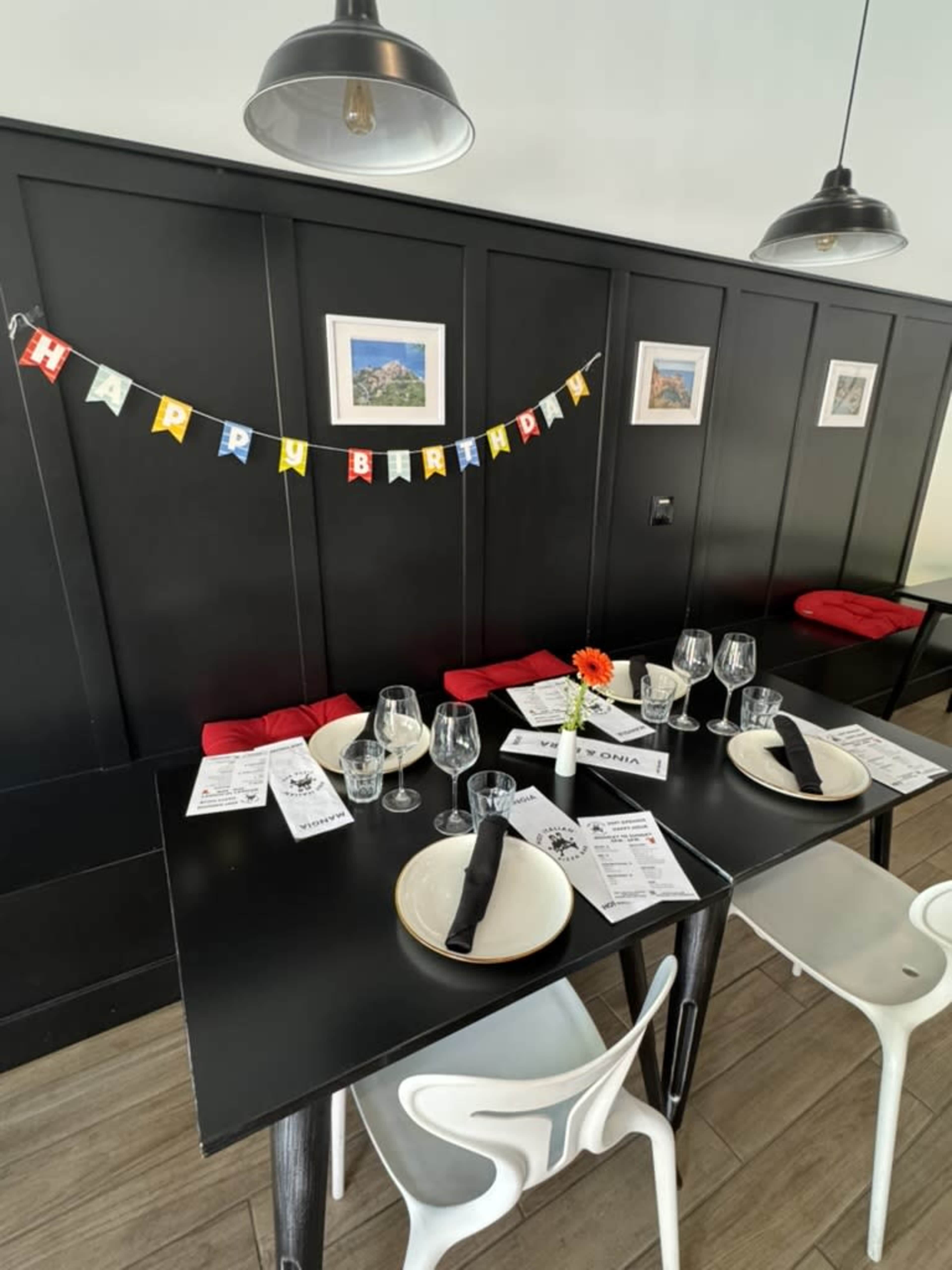 A birthday celebration setup features a black-walled dining area with a "Happy Birthday" banner, neatly arranged tables with menus, glasses, cutlery, and a decorative flower on a table.
