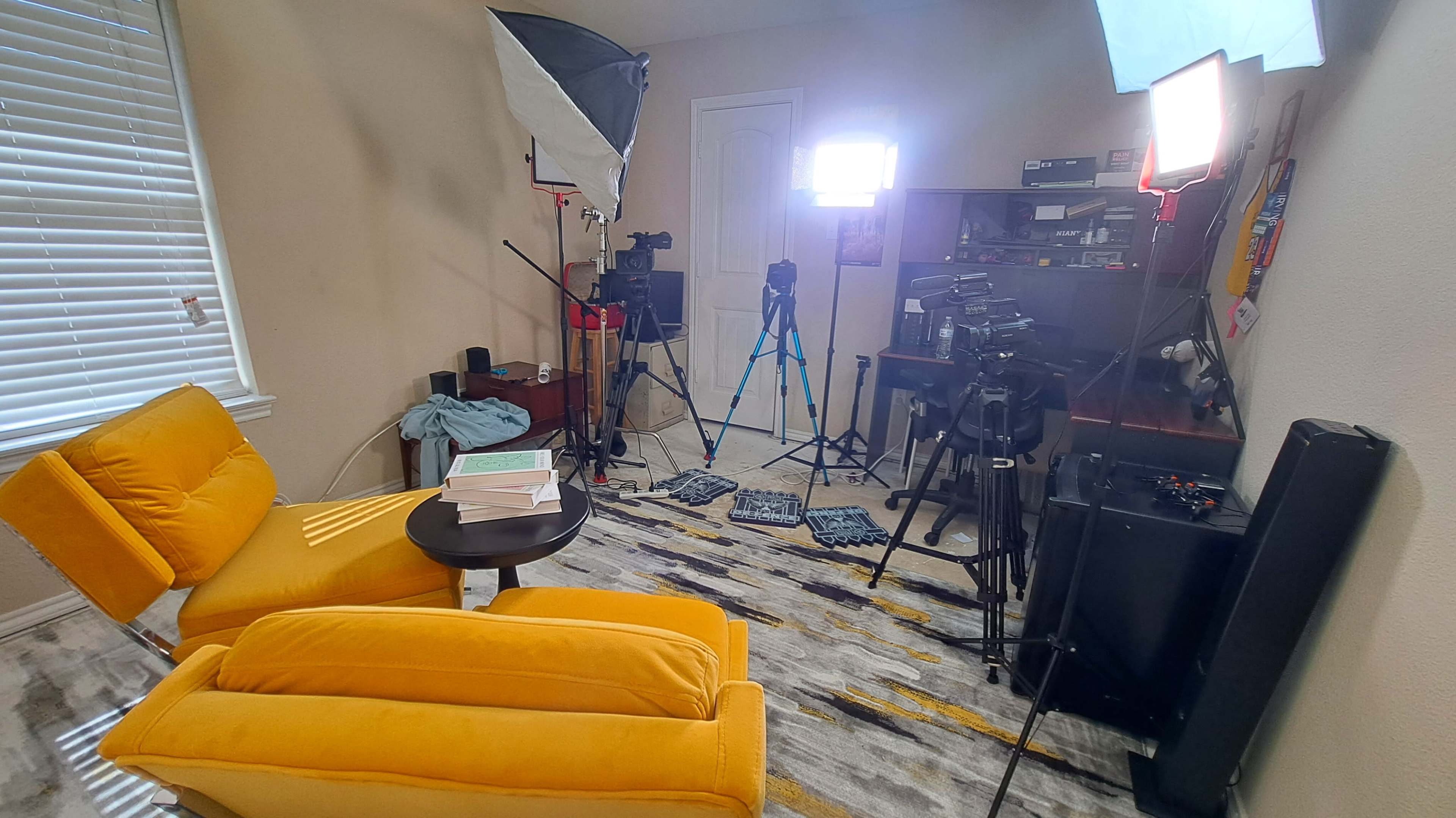 The image shows a brightly lit recording setup in a room featuring a yellow chair, a small table with scattered papers, and multiple cameras and tripods positioned around the space.