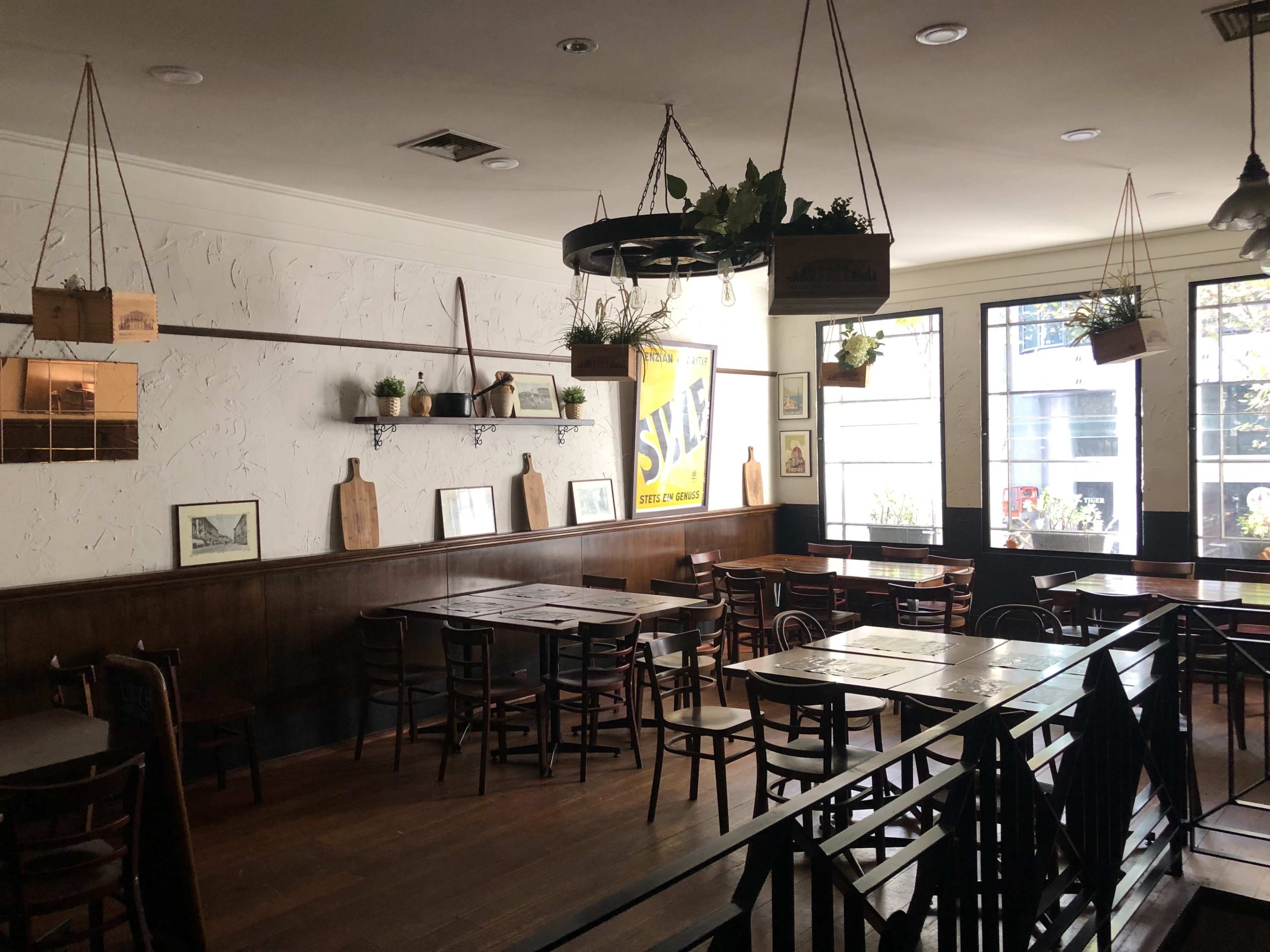 The image depicts a cozy restaurant with wooden tables and chairs, featuring plants hanging from the ceiling and a large wall-mounted sign.