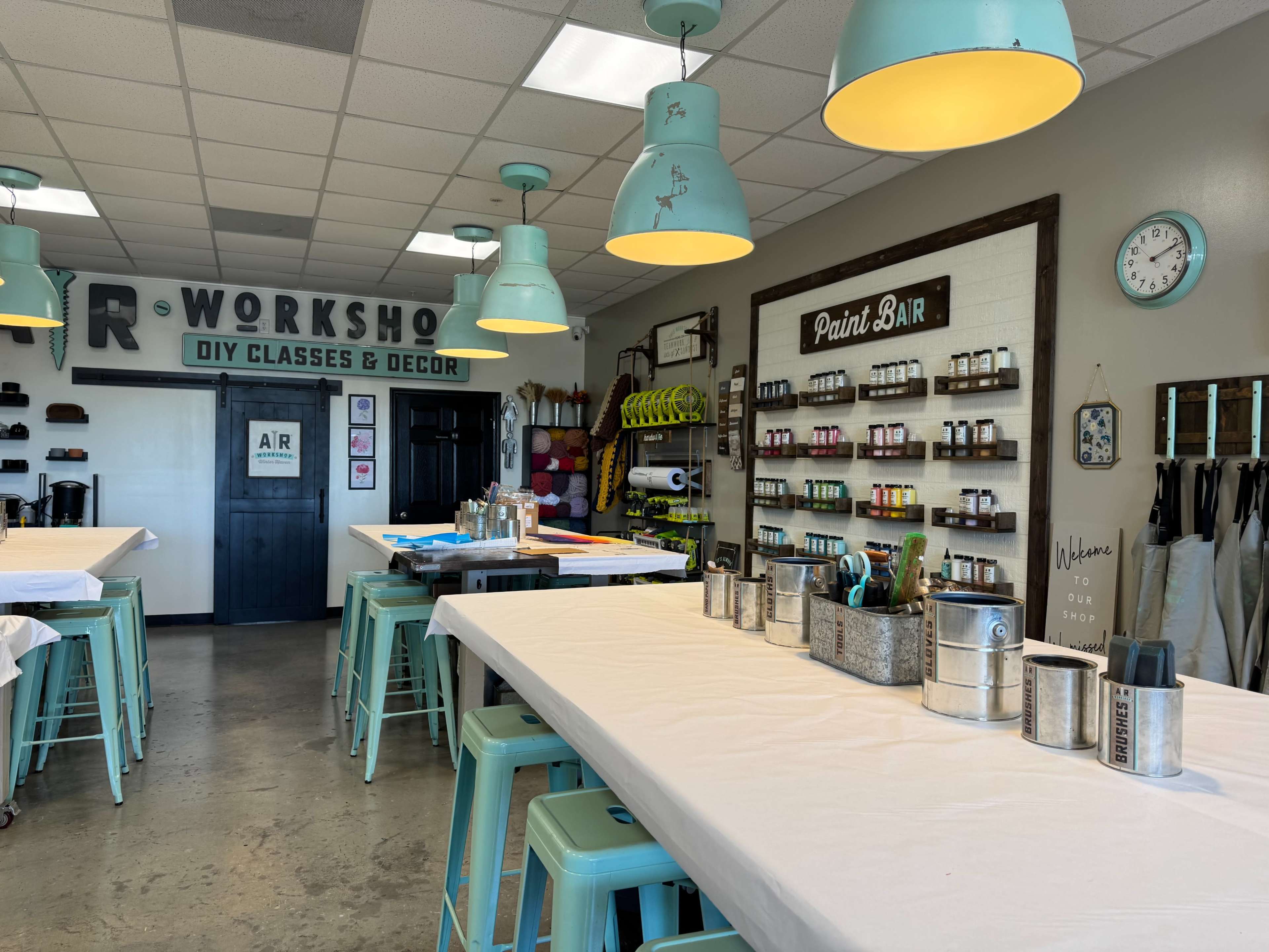 The image shows a brightly lit workshop space with several tables covered in white tablecloths, blue pendant lights hanging from the ceiling, and shelves displaying various art supplies.