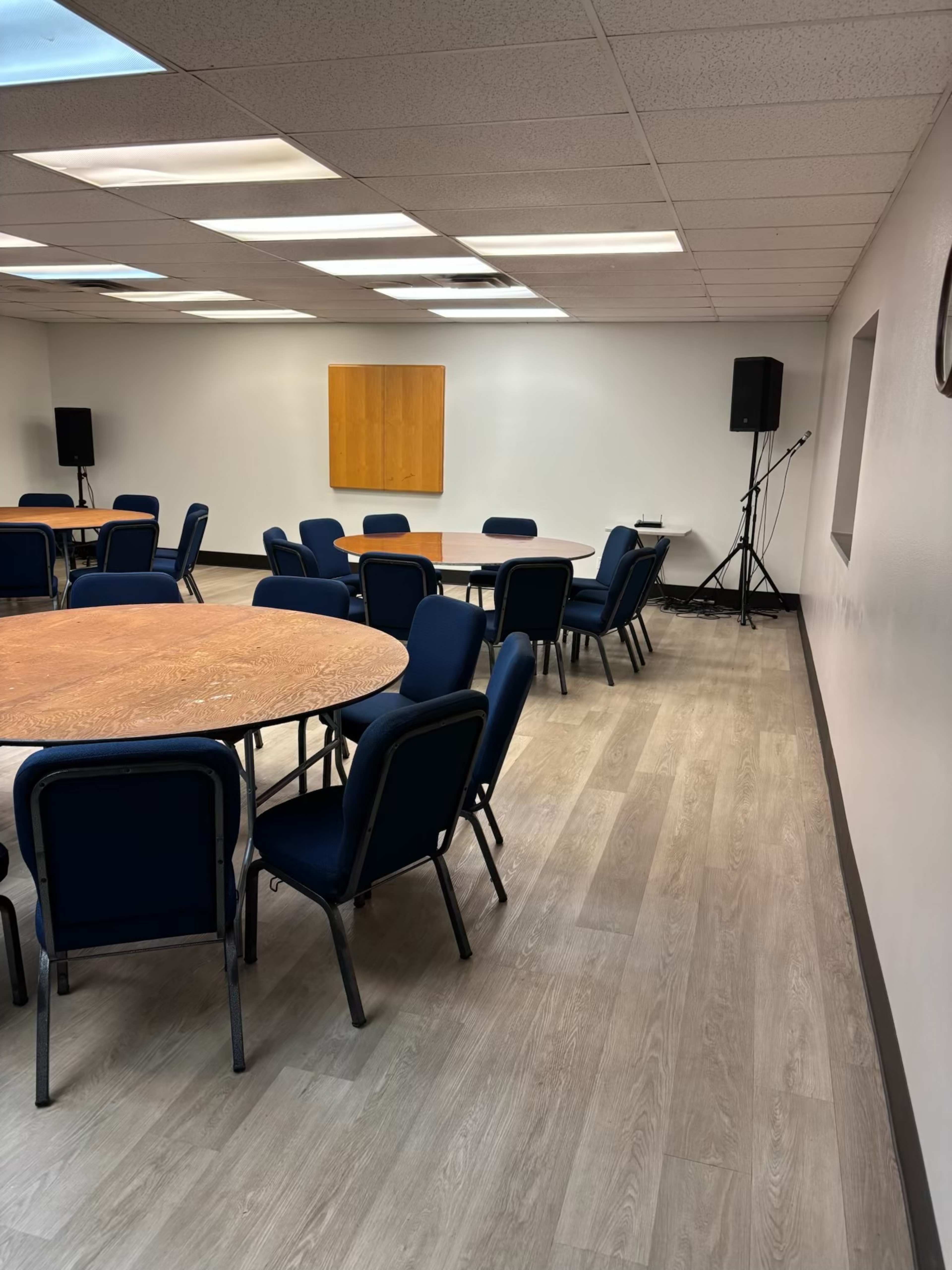 The image shows a meeting room with several round tables and navy blue chairs arranged neatly, along with a speaker setup in one corner.