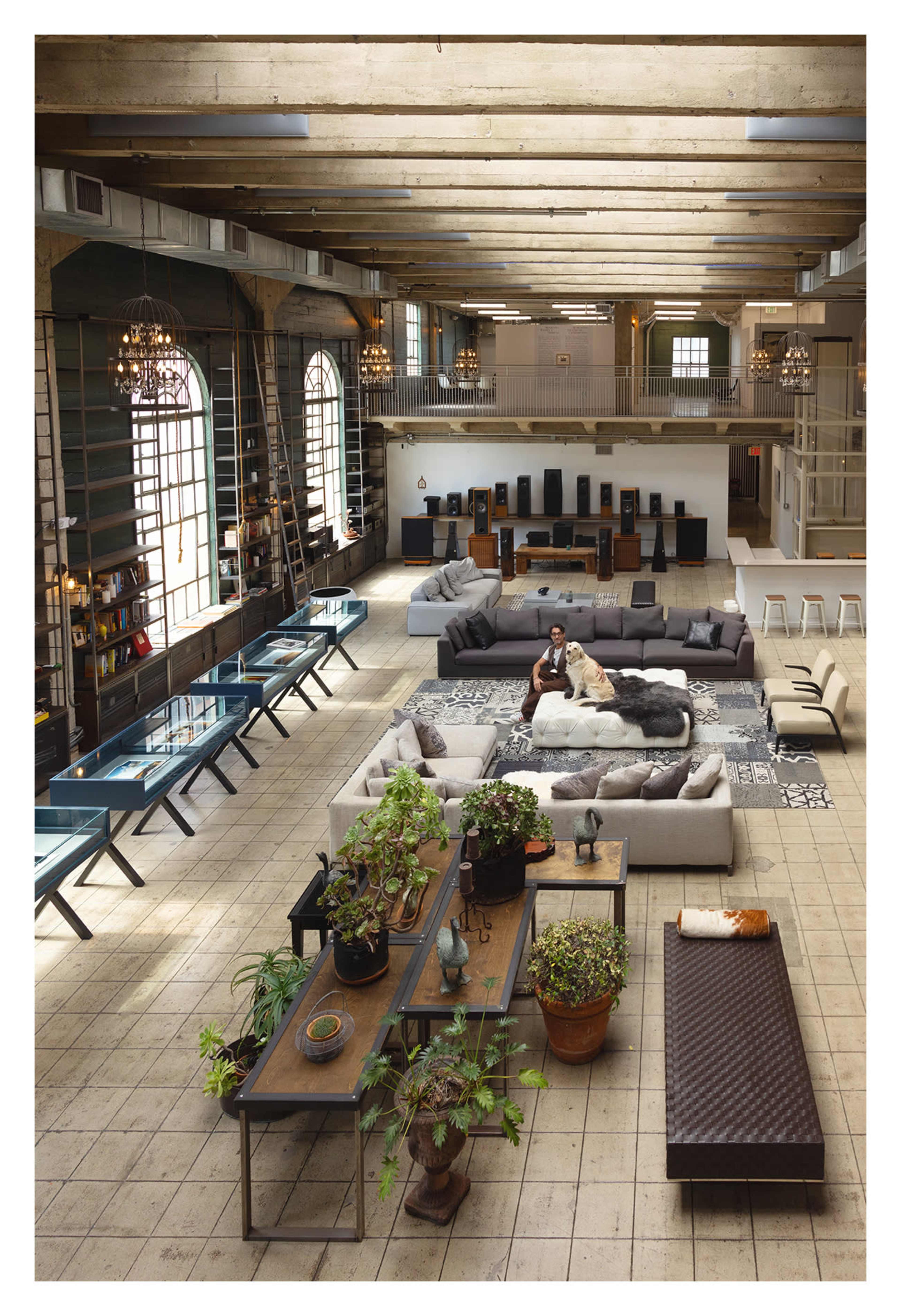 The image shows a spacious industrial-style loft with high ceilings, large windows, multiple seating areas, and various plants arranged on long tables.