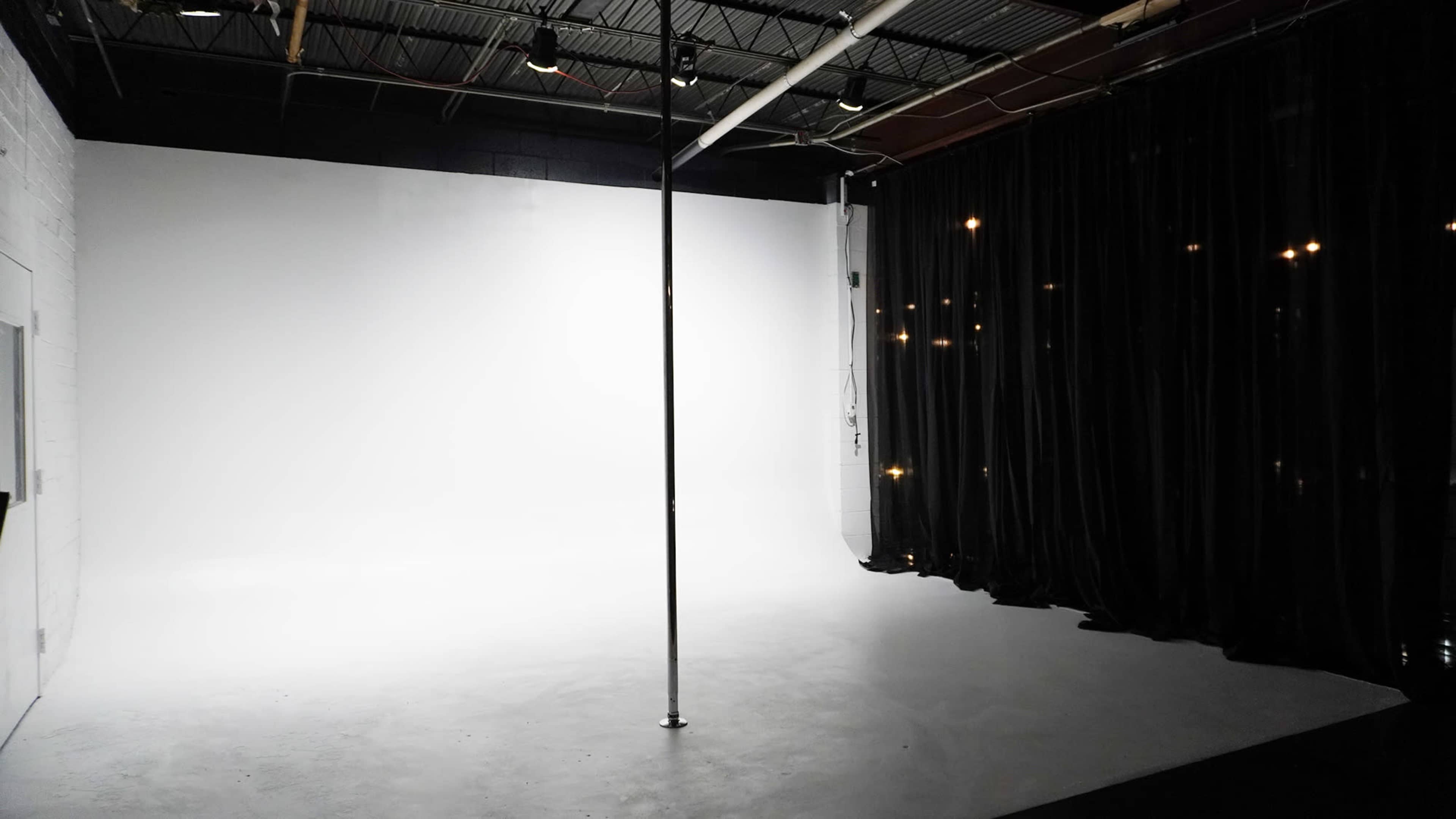 The image shows a spacious, empty studio with a vertical pole and a black curtain against a white backdrop.