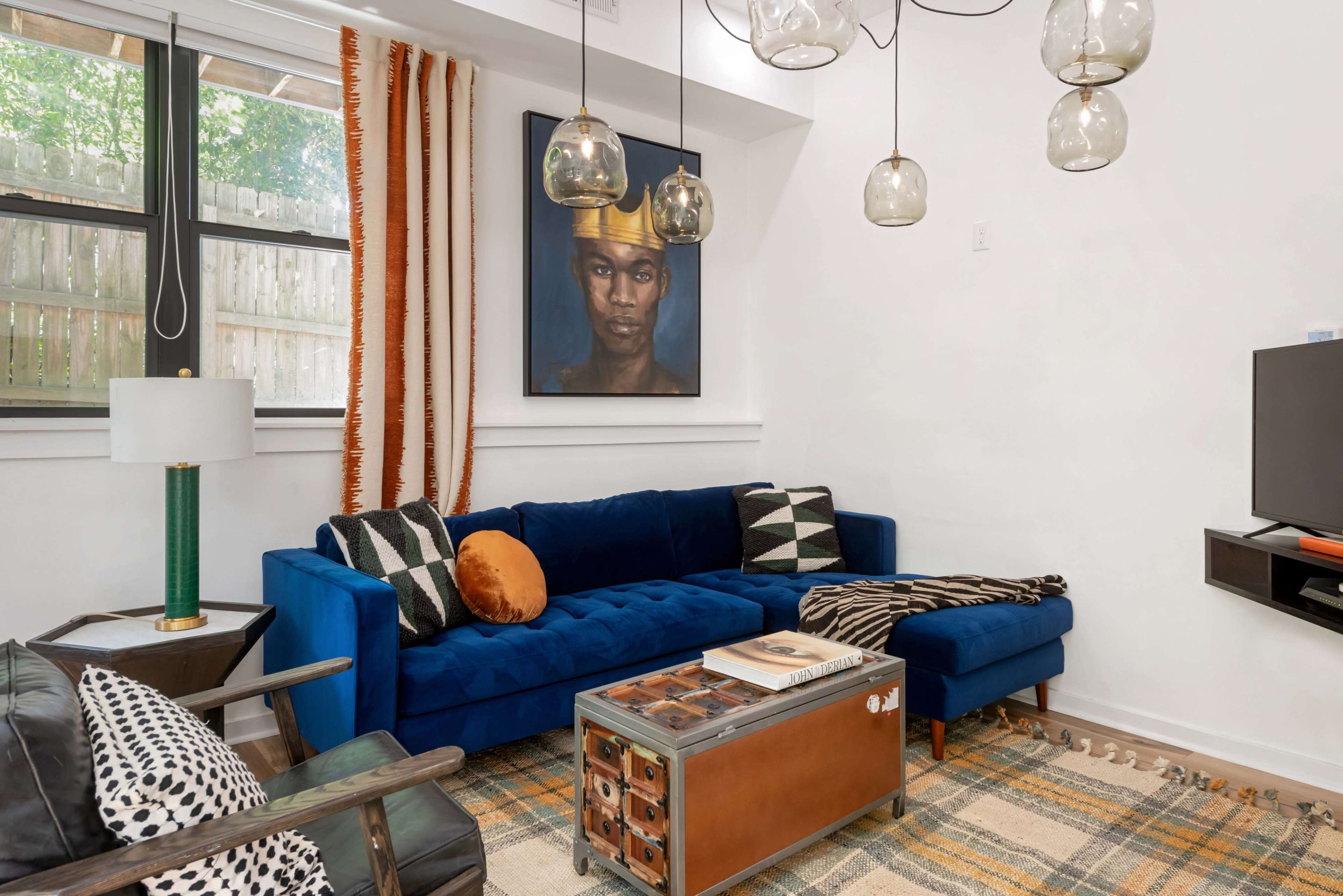 The image shows a modern living room featuring a blue sectional sofa, a wooden coffee table, a patterned rug, and pendant lighting, with a large framed portrait hanging on the wall.