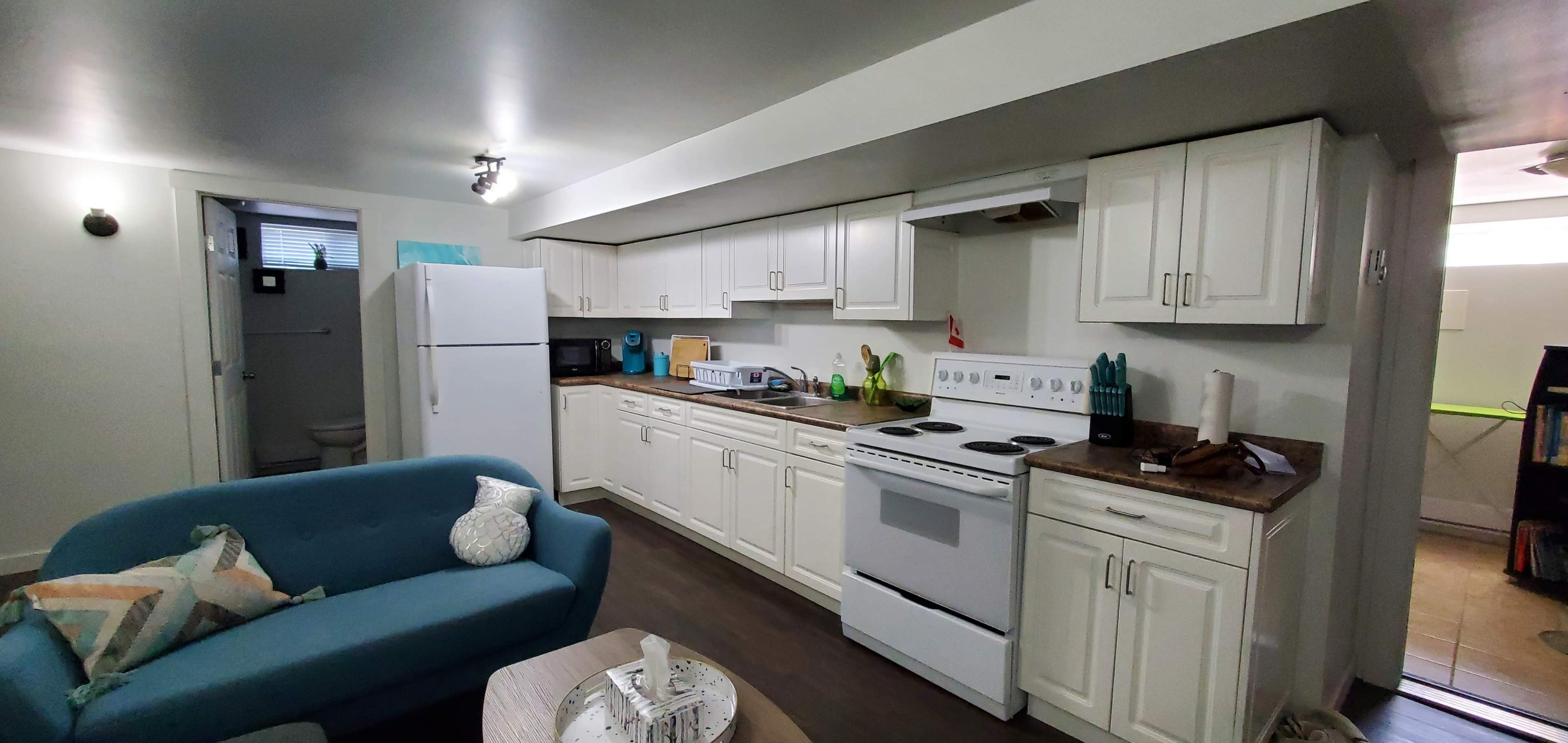 The image shows a modern kitchen with white cabinetry, a stove, a fridge, and a small seating area with a blue couch.