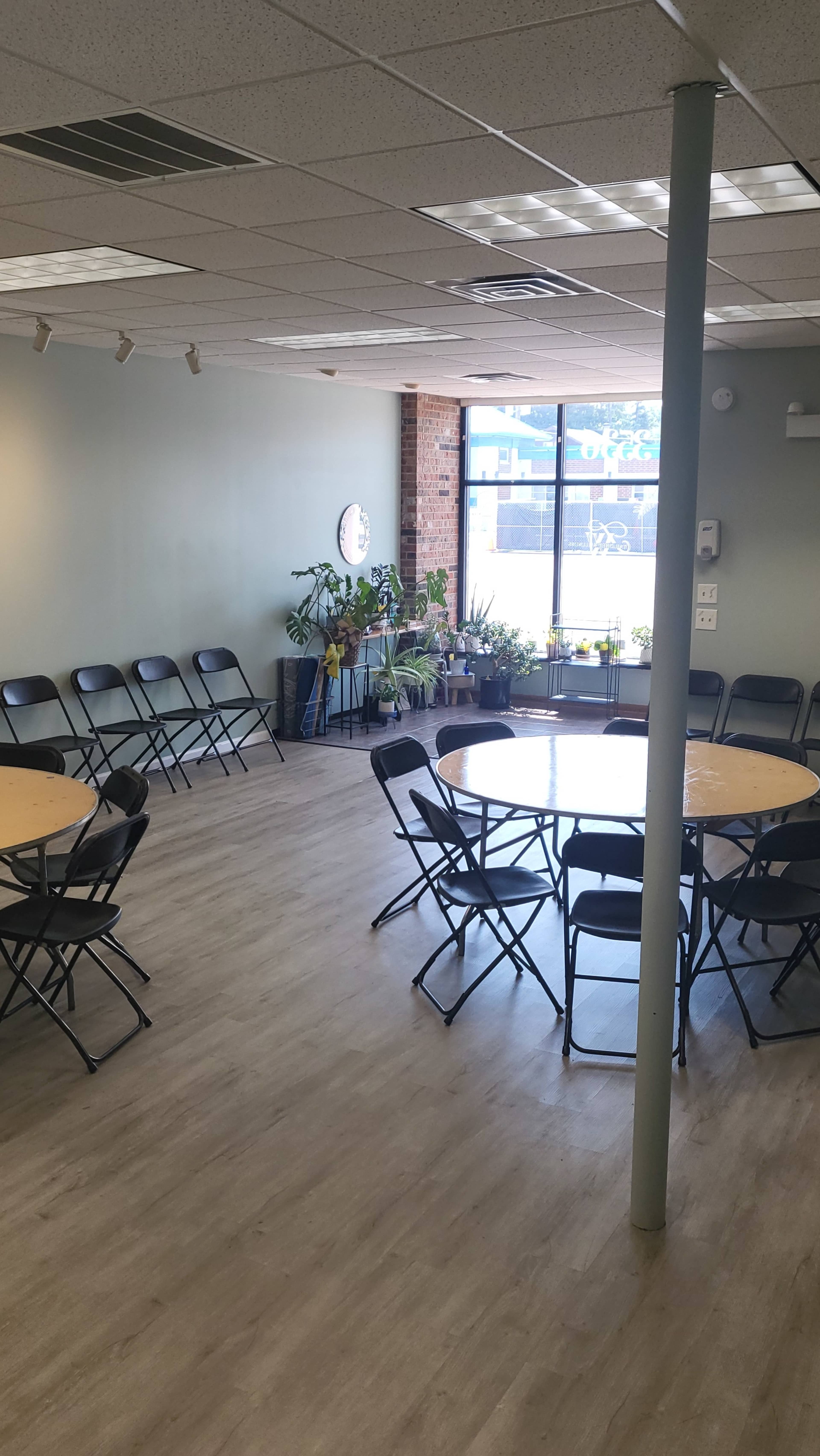 A spacious room with multiple round tables and black folding chairs arranged around them, featuring large windows that allow natural light and potted plants placed throughout the space.