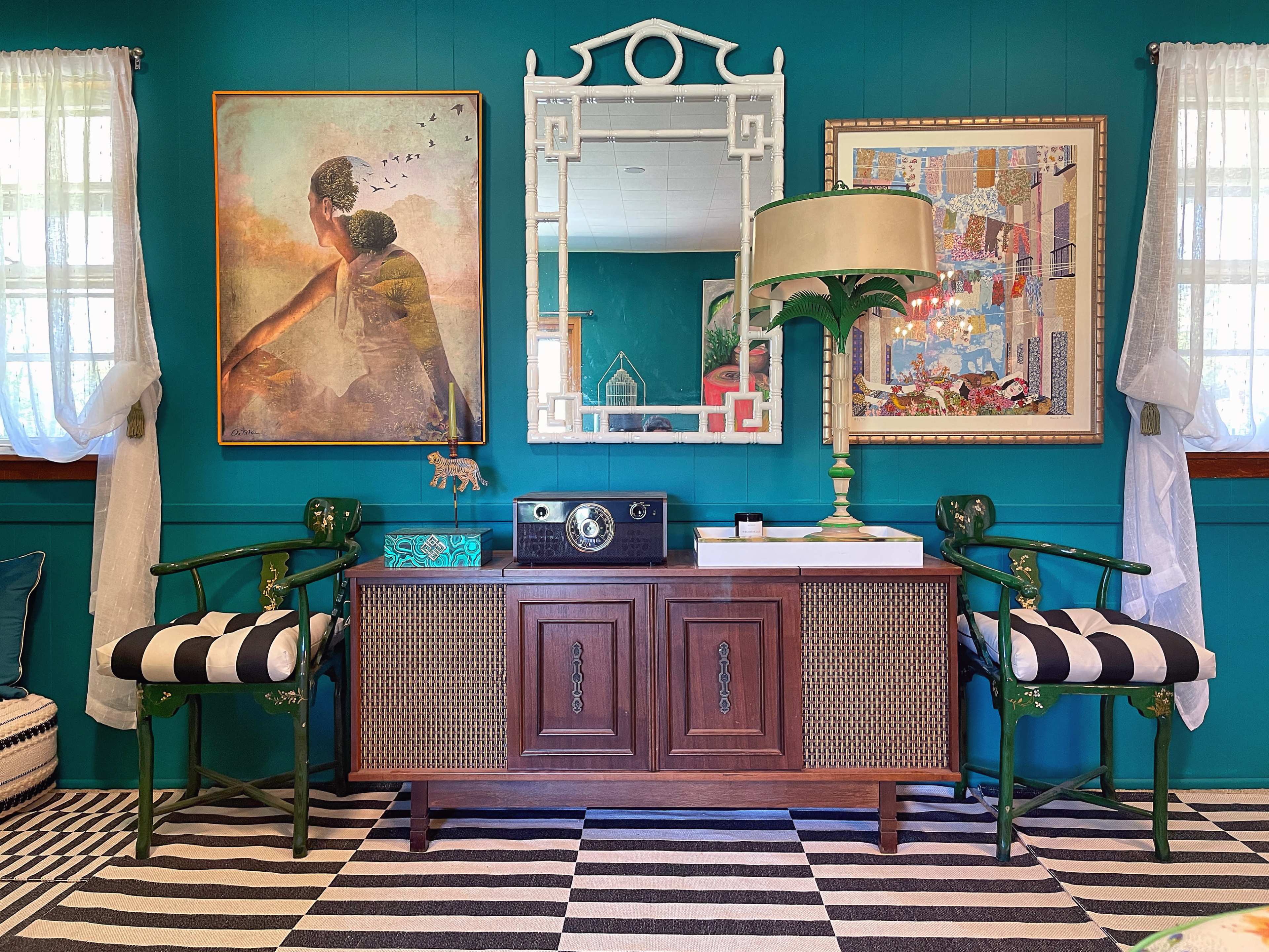 A wooden console table with patterned decor and two green chairs is placed against a teal wall, flanked by framed artwork and a mirror.