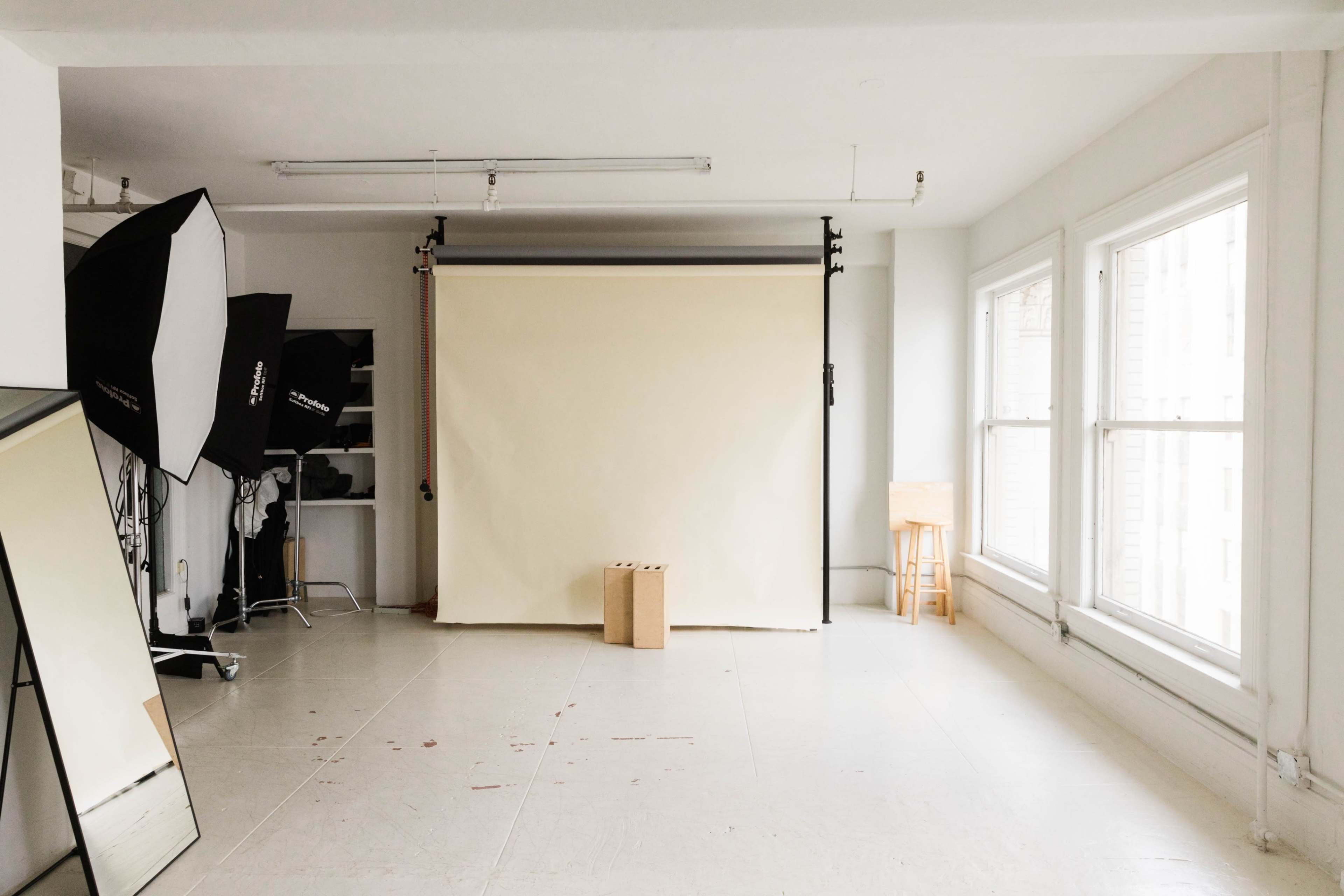 A photography studio features a beige backdrop, two large softbox lights, a mirror, and a wooden stool situated in a well-lit space with large windows.