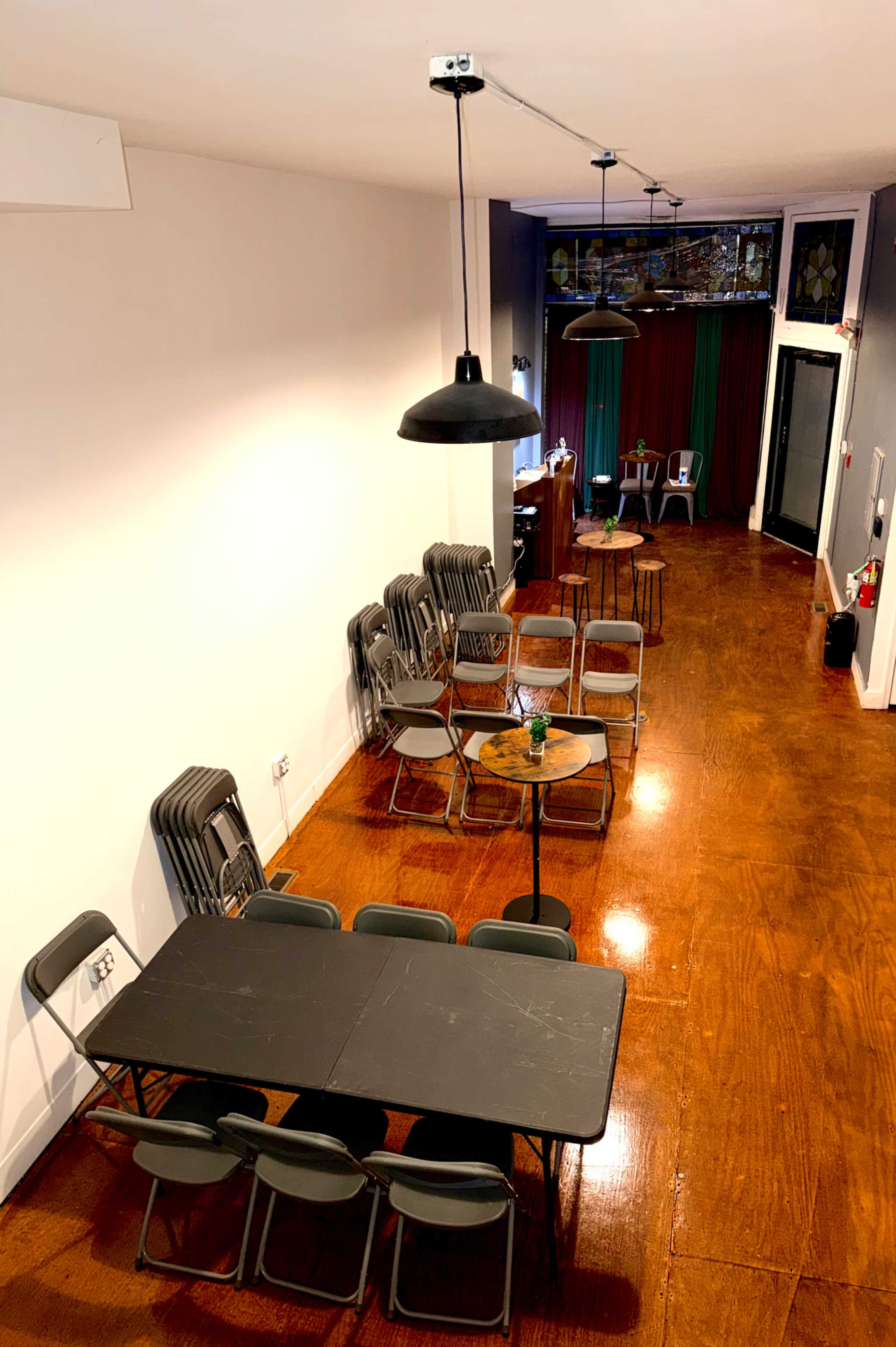 The image shows a spacious room with stacked chairs along one side, several tables in the center, and hanging lamps above.