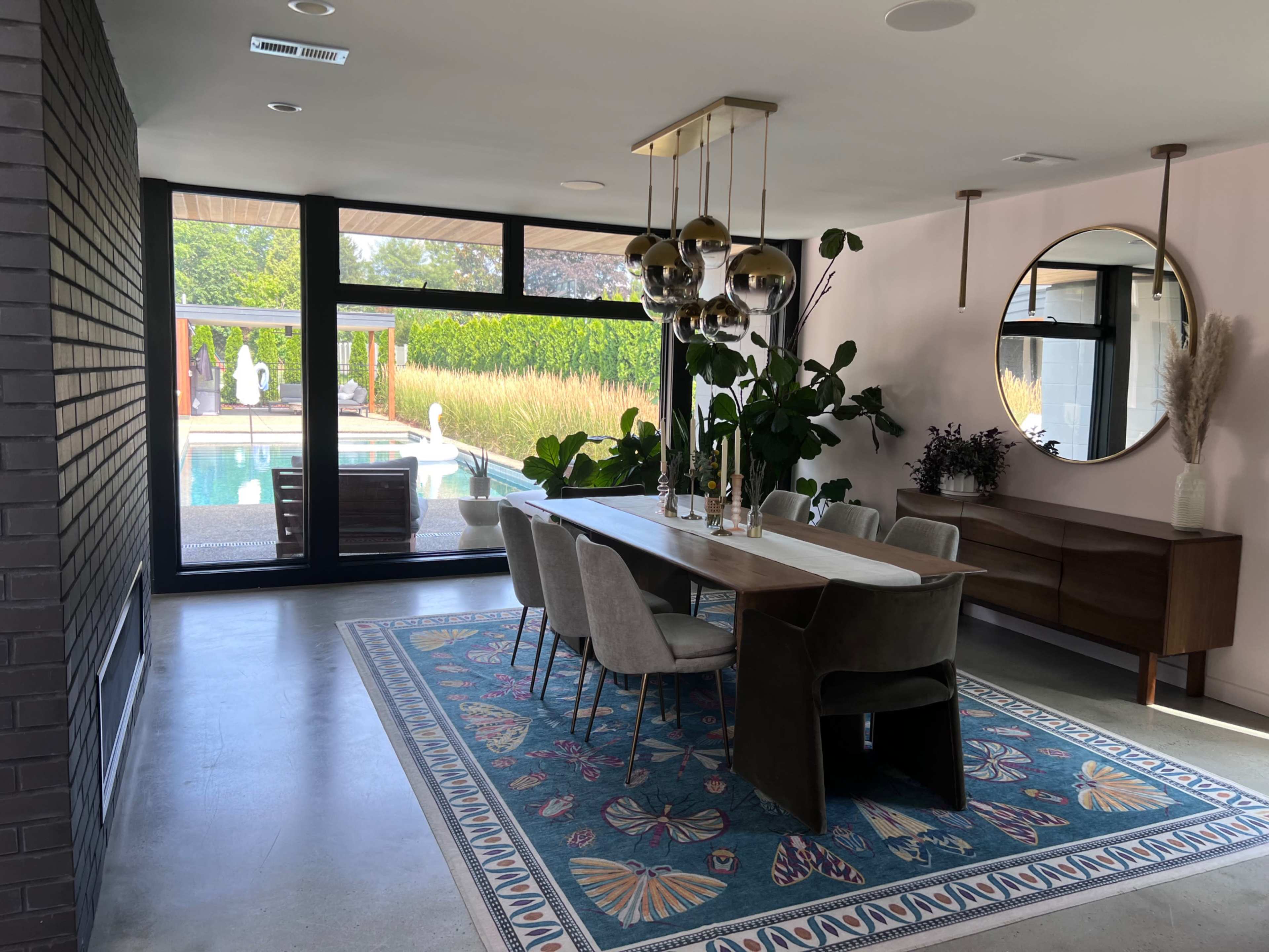 A dining area features a long table with six chairs, a large mirror, and plants, overlooking a pool through floor-to-ceiling windows.