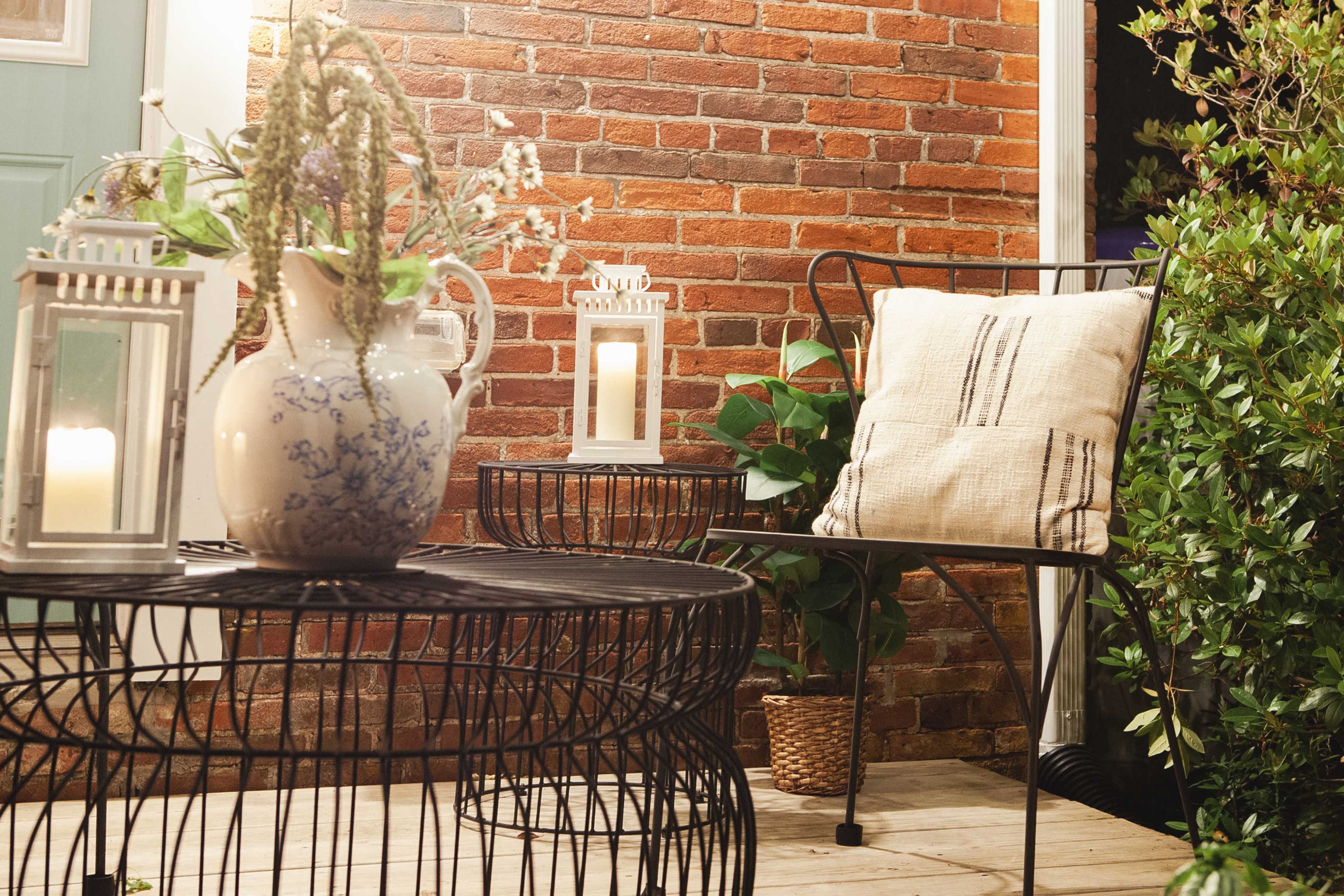 A cozy outdoor seating area features a black wire table, a comfortable cushion chair, a vase with flowers, and lanterns against a brick wall backdrop.