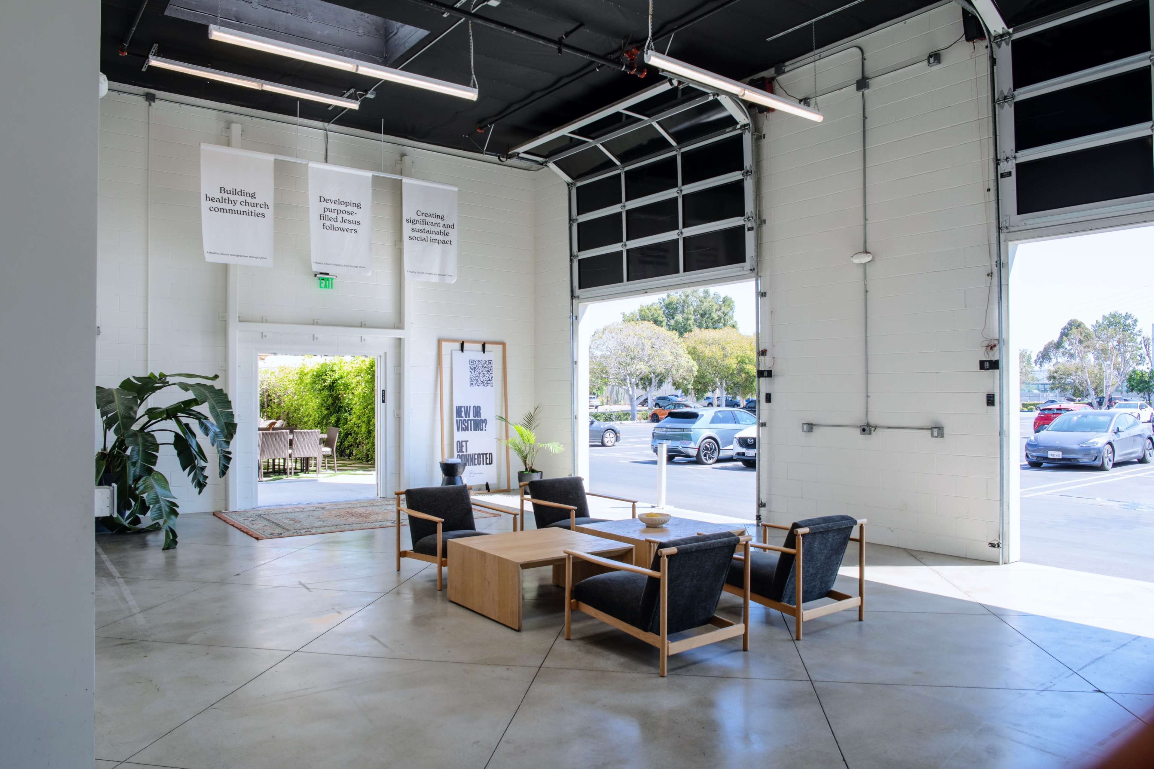 The image shows a modern lobby area with a few wooden and upholstered chairs arranged around a table, and large garage doors leading to an outdoor space.