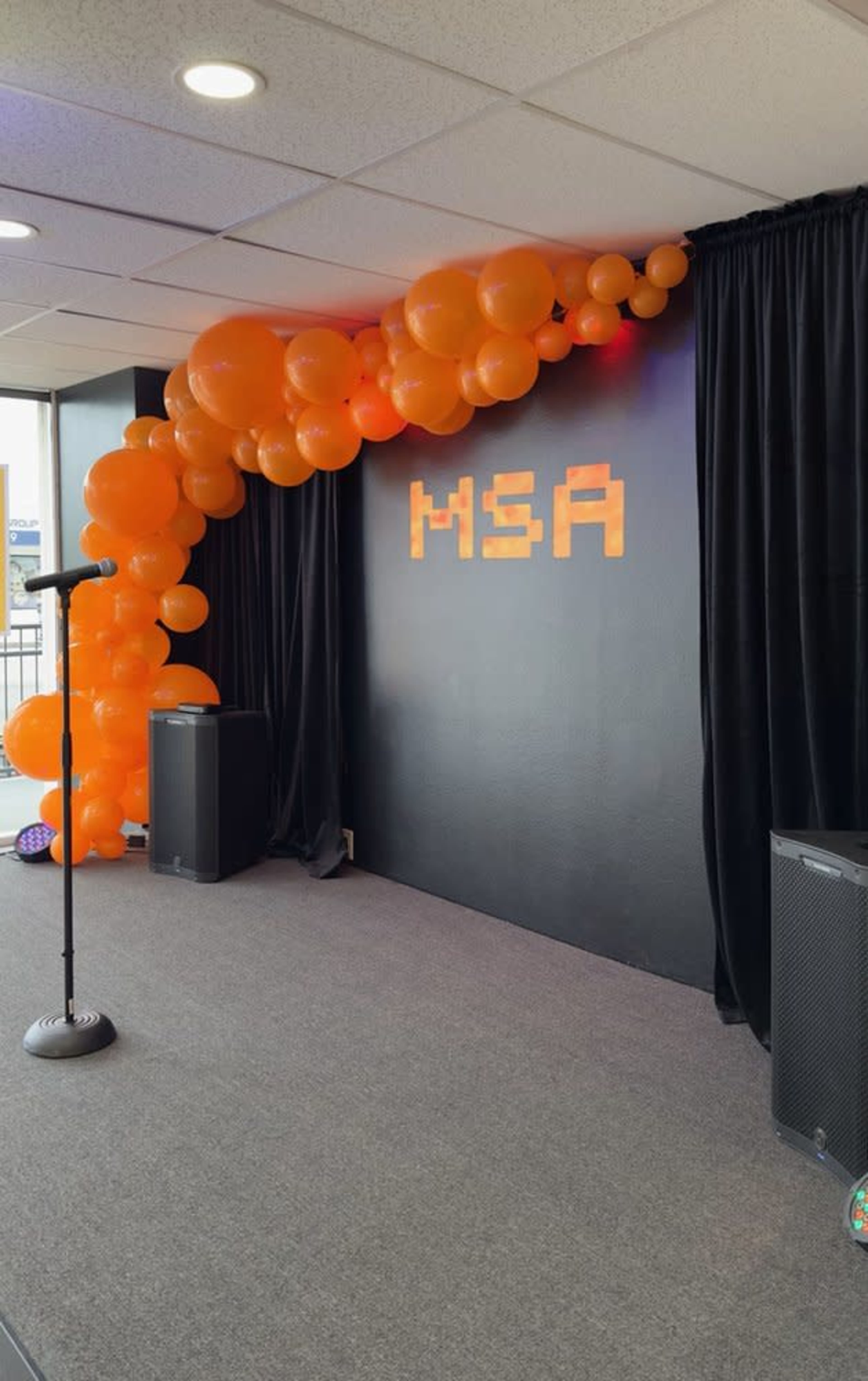 The image shows a stage decorated with a balloon arch in shades of orange and the letters "MSA" displayed on a black backdrop.