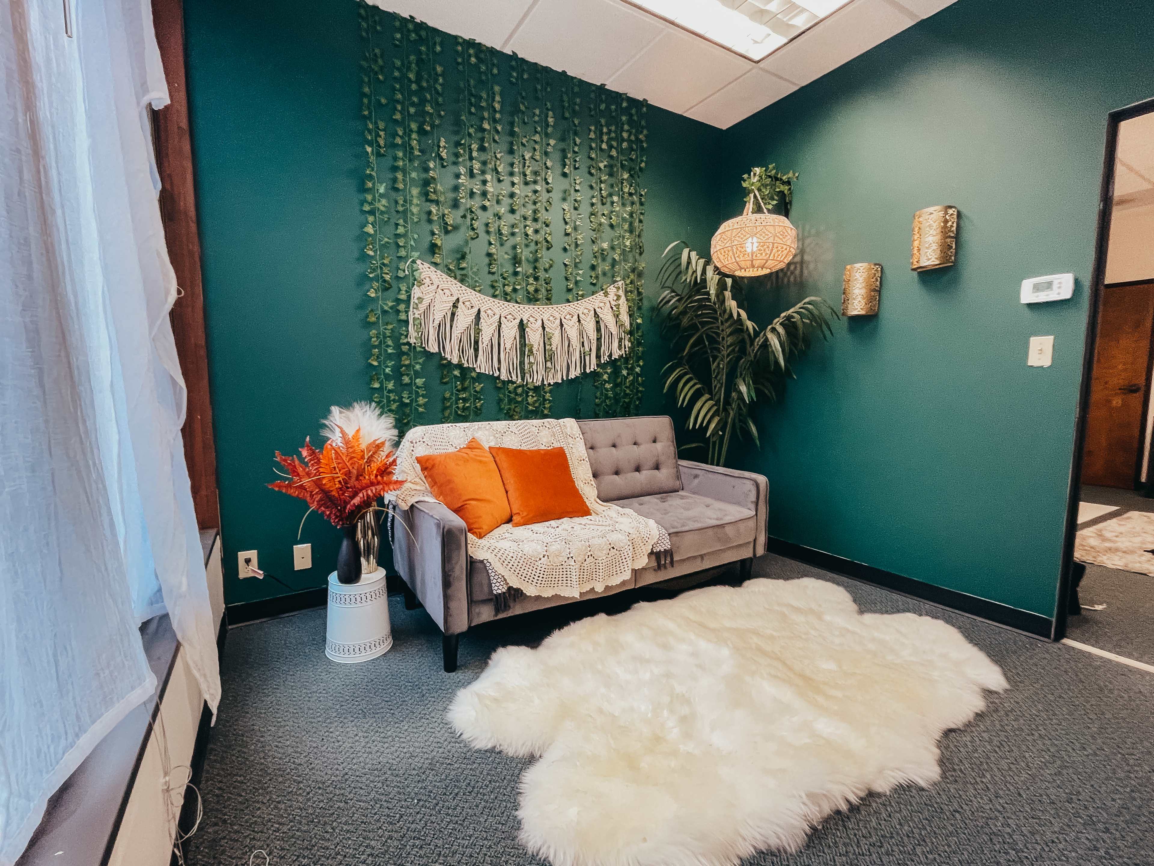 The image depicts a cozy room featuring a gray couch adorned with orange pillows, a decorative wall hanging, and greenery, alongside a white faux fur rug on a carpeted floor.