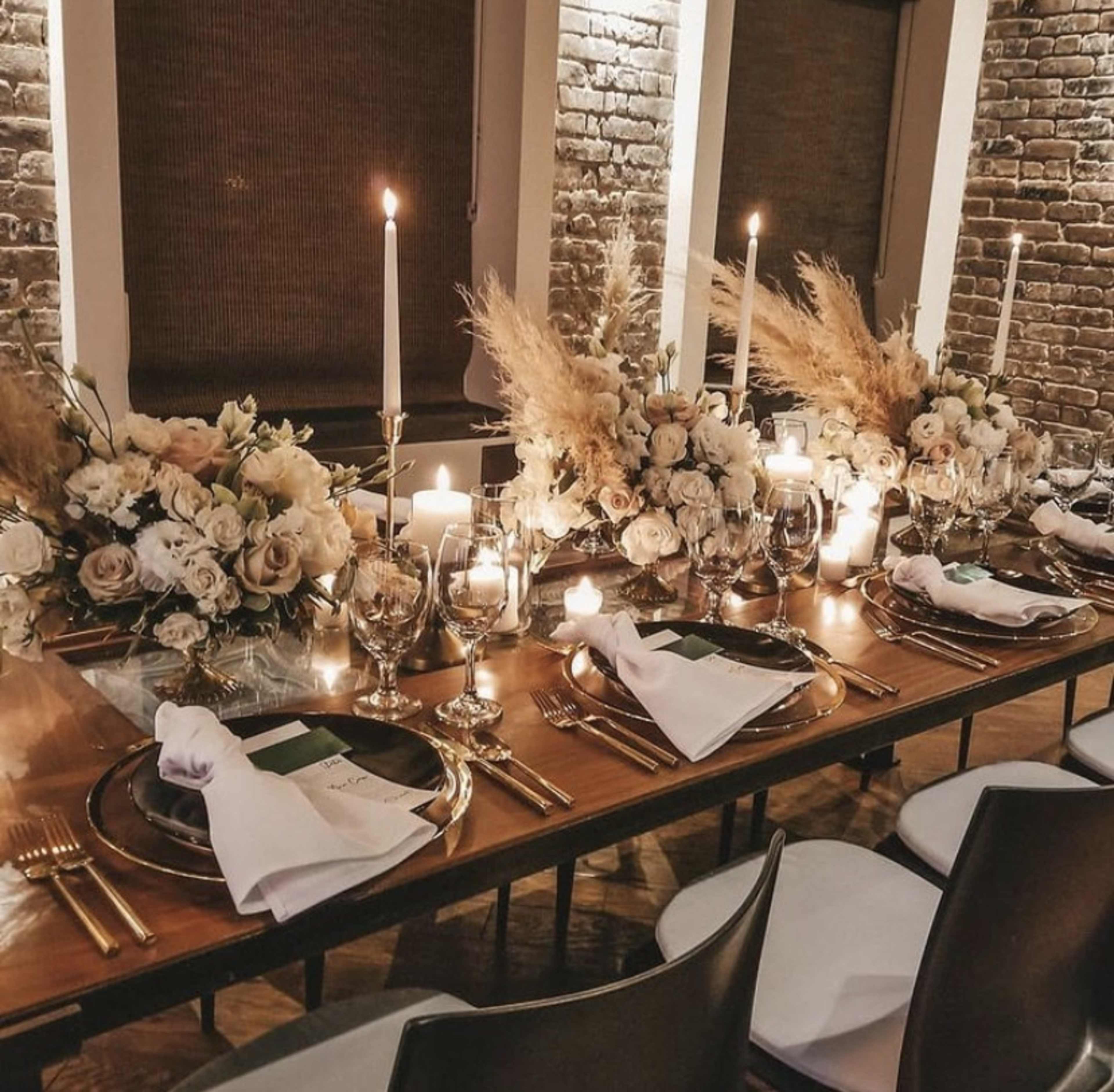 A long dining table is set with elegant centerpieces, candles, and glassware in a room with exposed brick walls.