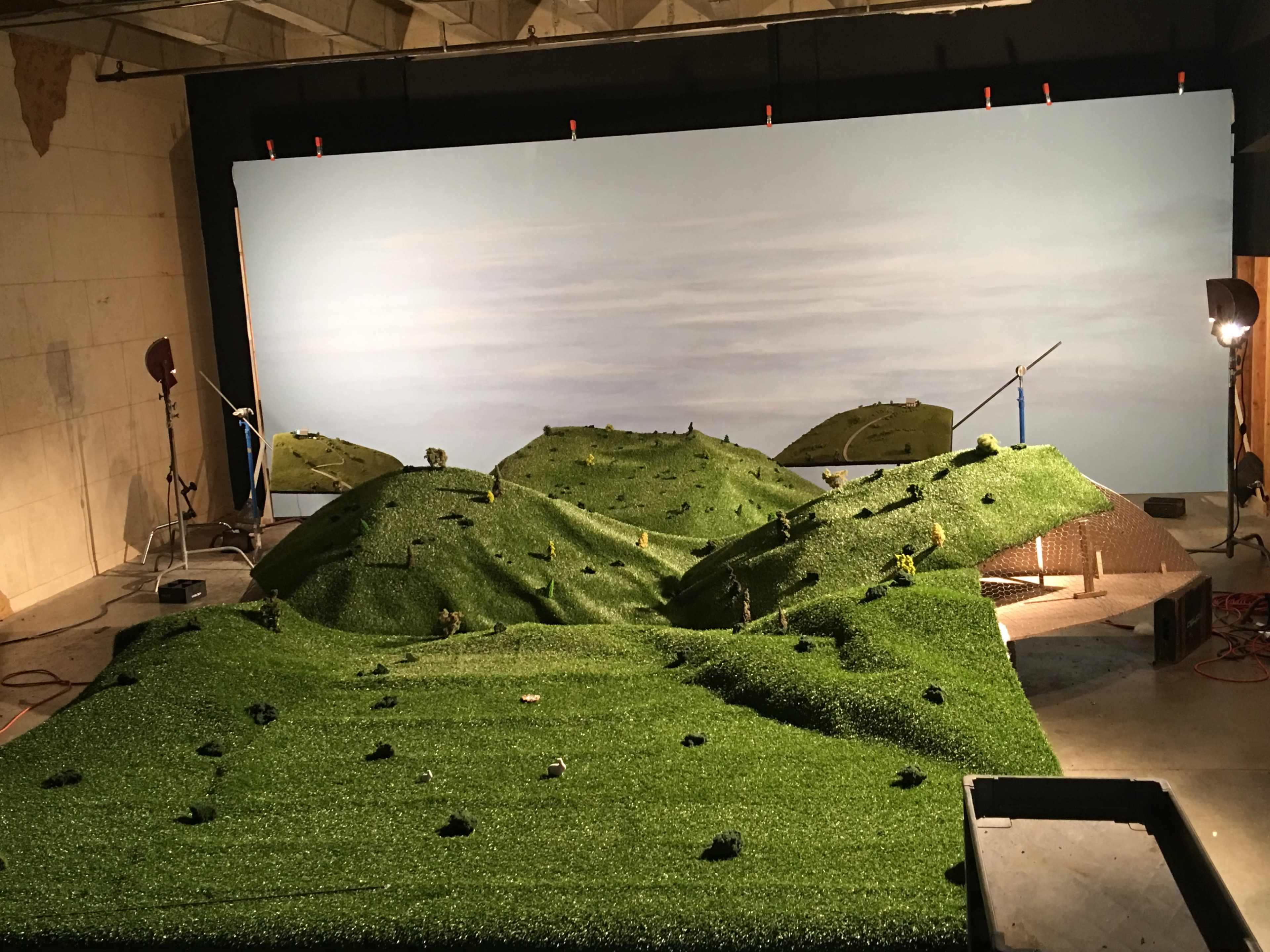 The image shows a scale model of a hilly landscape with artificial grass, set against a backdrop of a cloudy sky.