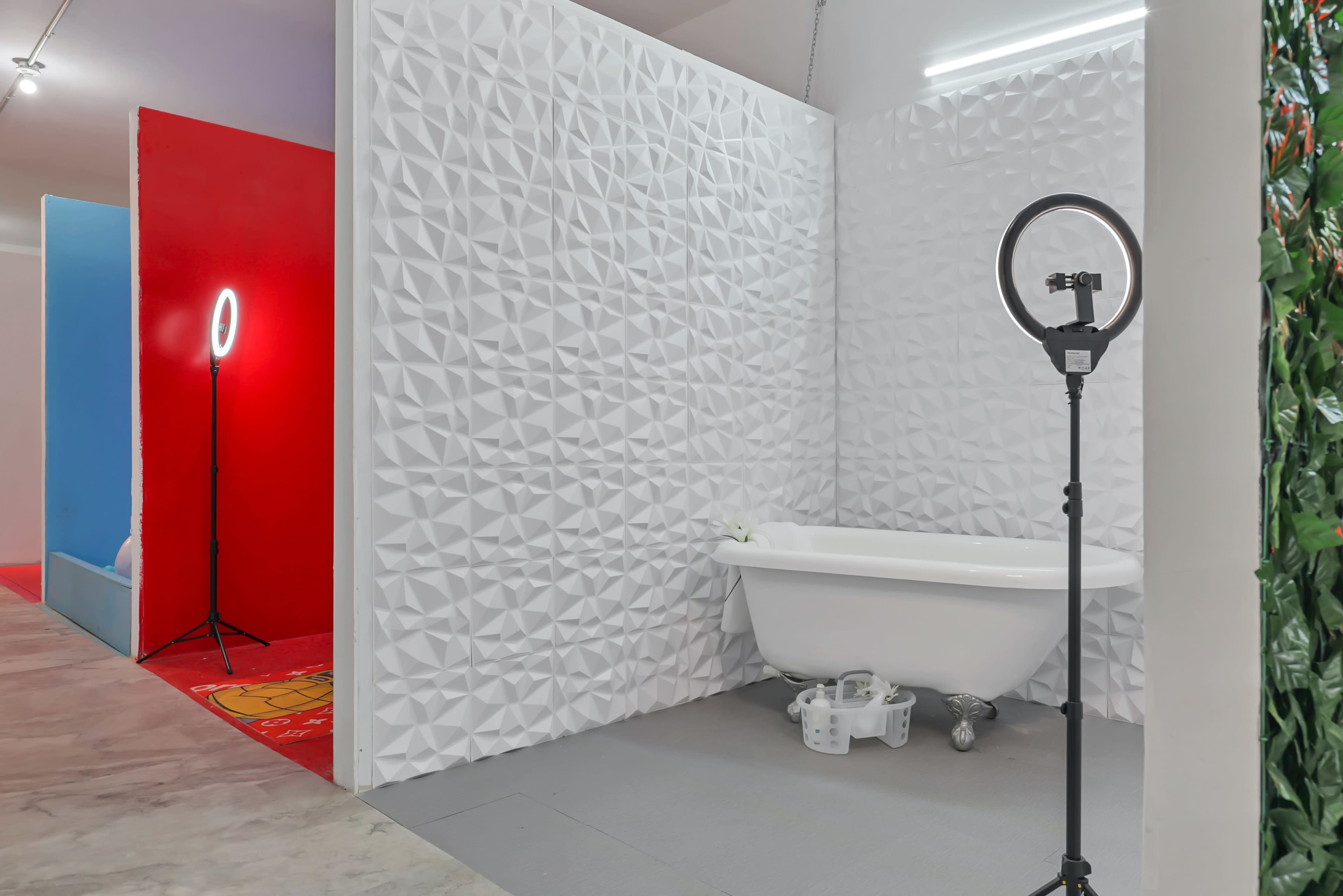 A bright bathroom setup with a freestanding bathtub against a textured white wall, accompanied by a ring light on a tripod.