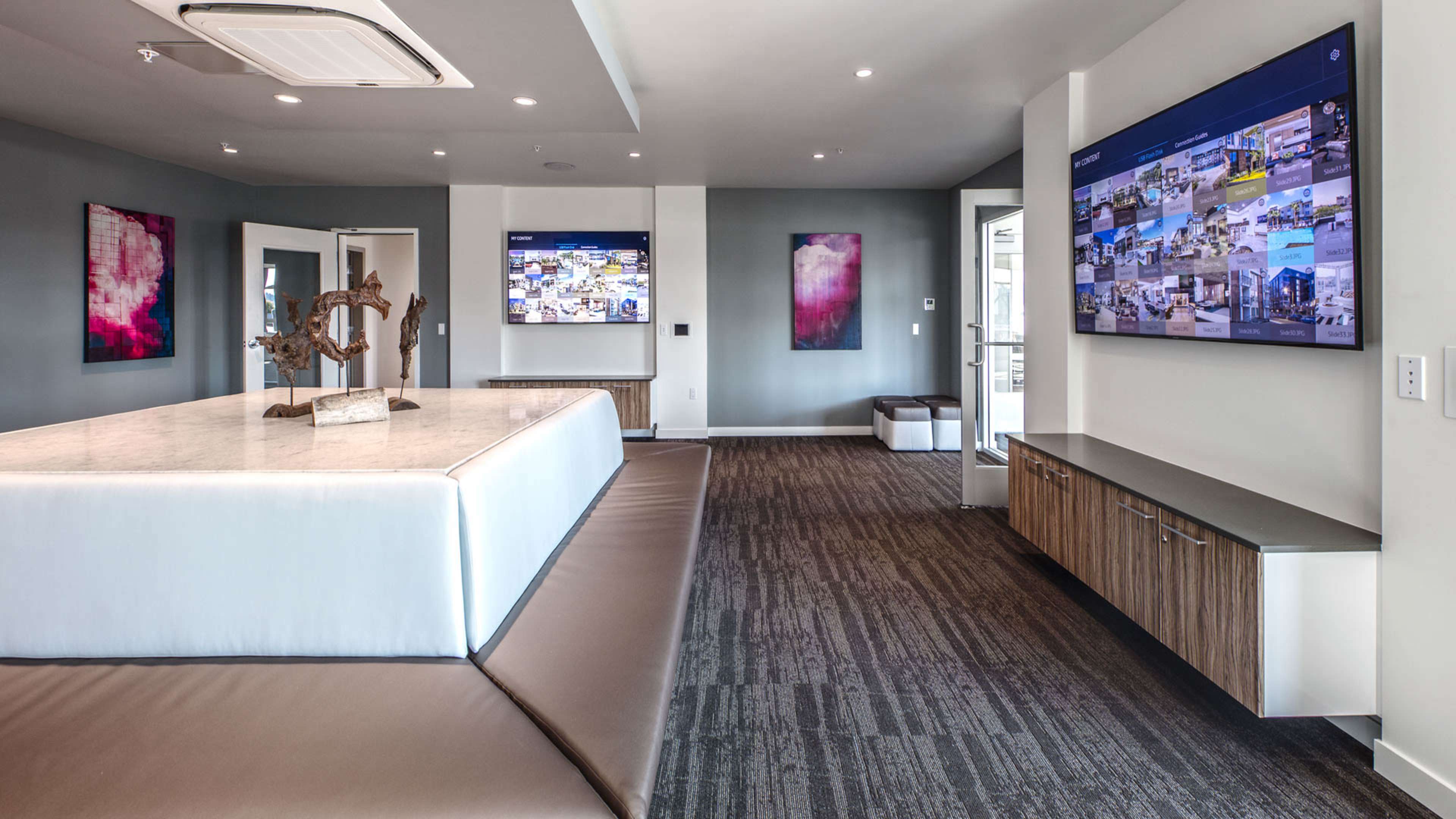 A modern interior space featuring a large central island with a marble countertop, two wall-mounted screens displaying images, and contemporary artwork on the walls.