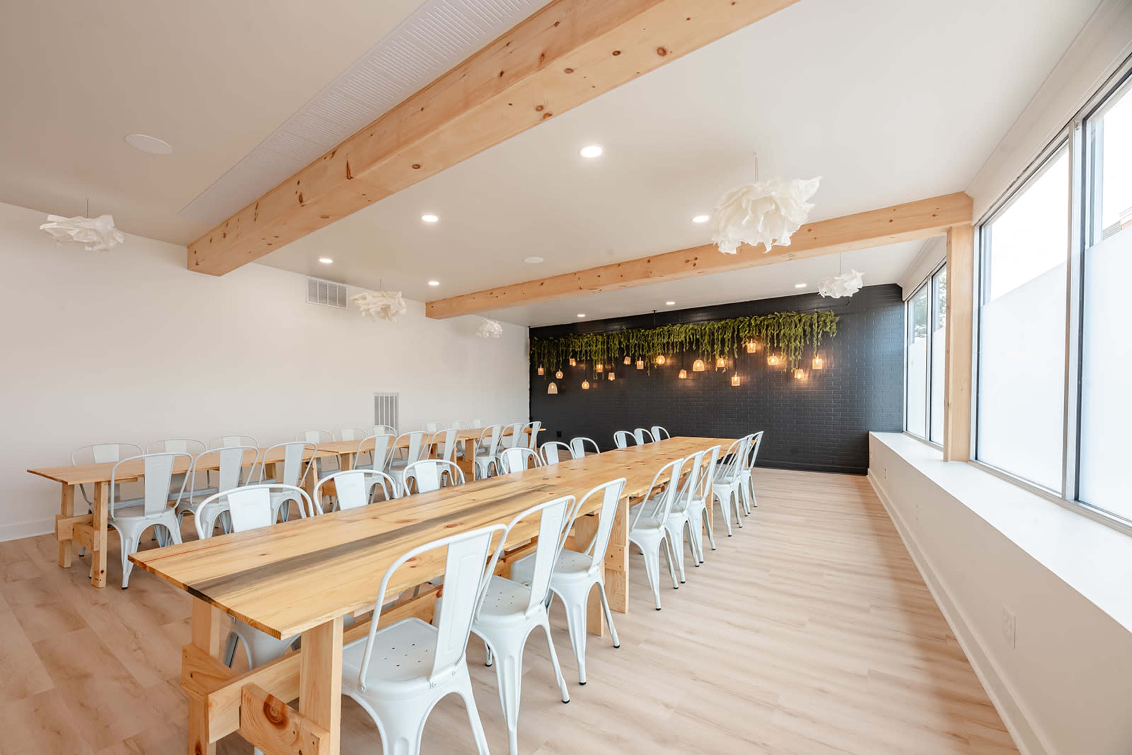 A long wooden table with metal chairs is set in a spacious, well-lit room featuring decorative hanging lights and greenery on the wall.