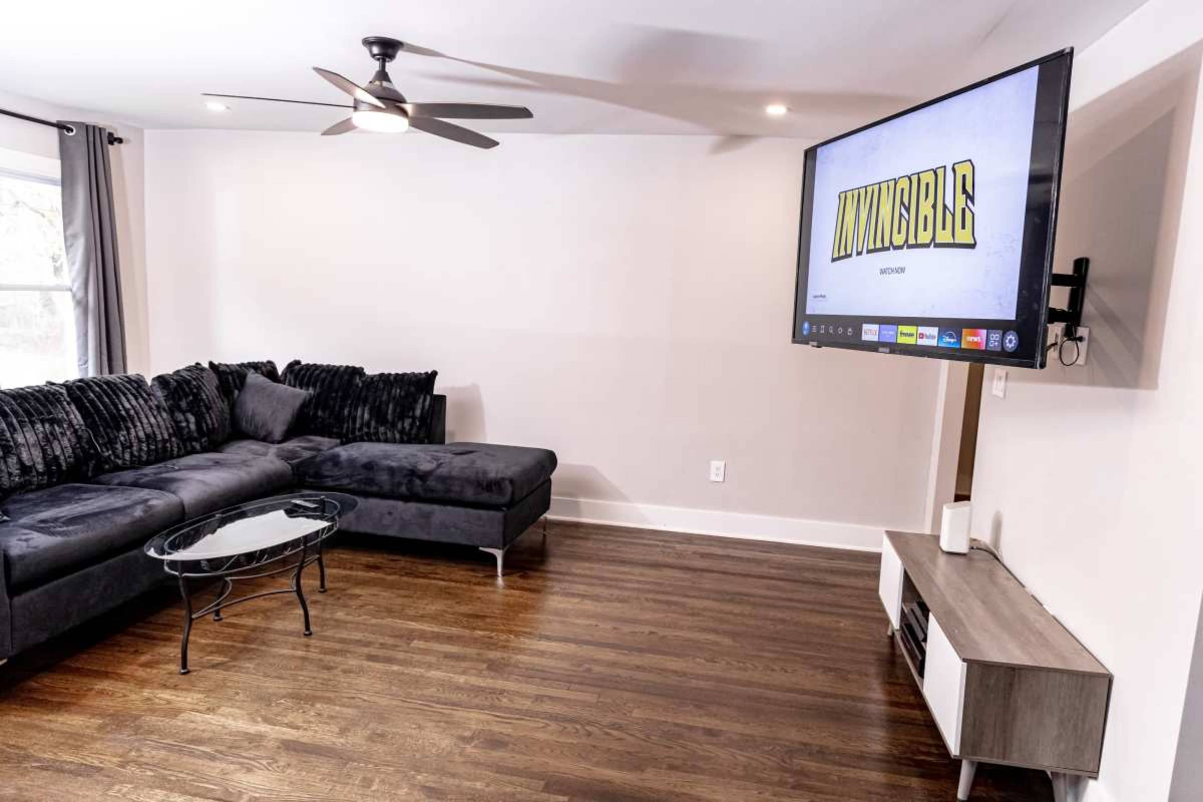A modern living room features a dark sectional sofa, a glass coffee table, and a wall-mounted television displaying the title "Invincible."