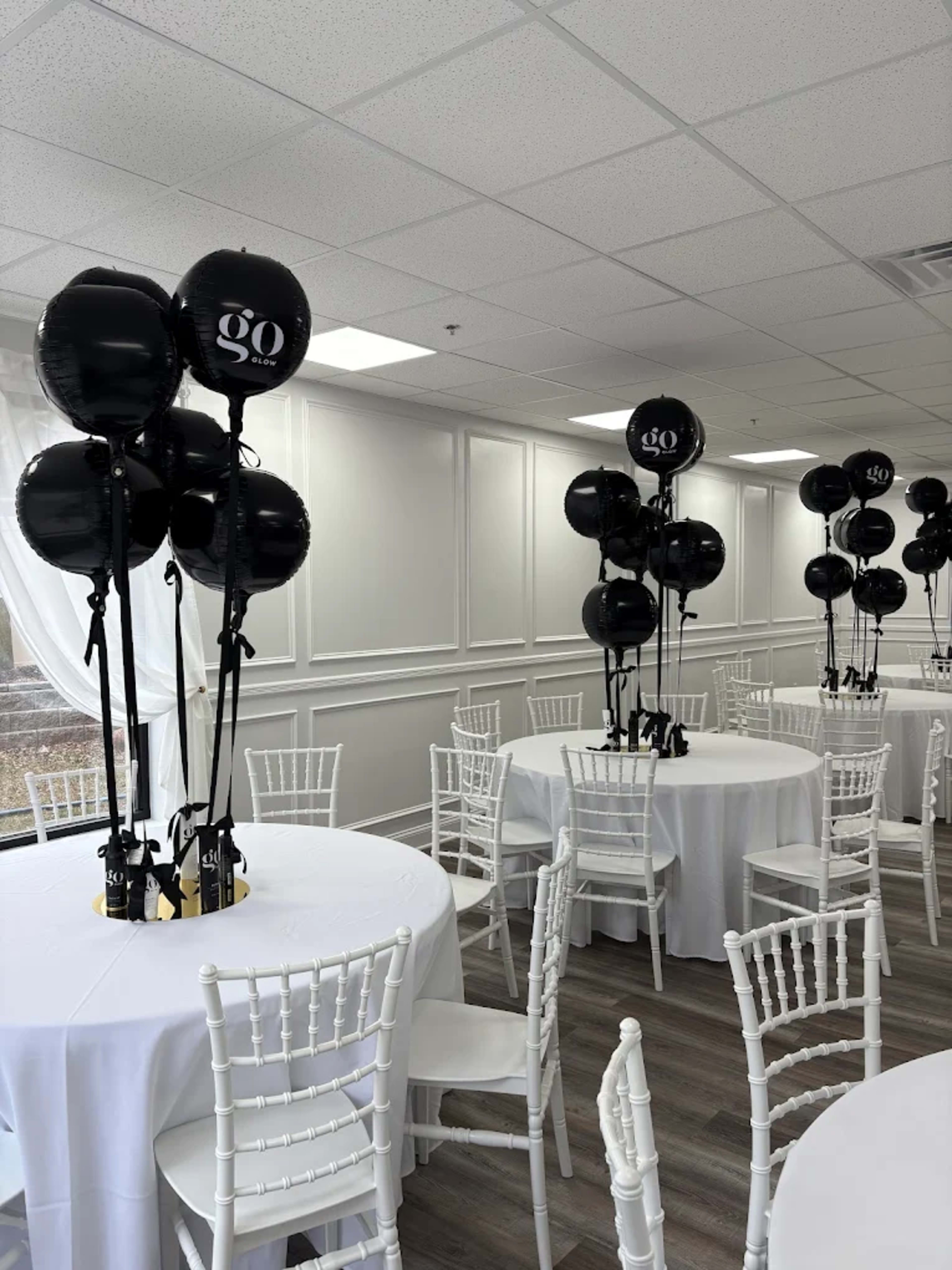 The image shows a banquet room with white tables set for an event, decorated with black balloons standing on tall stands at each table.