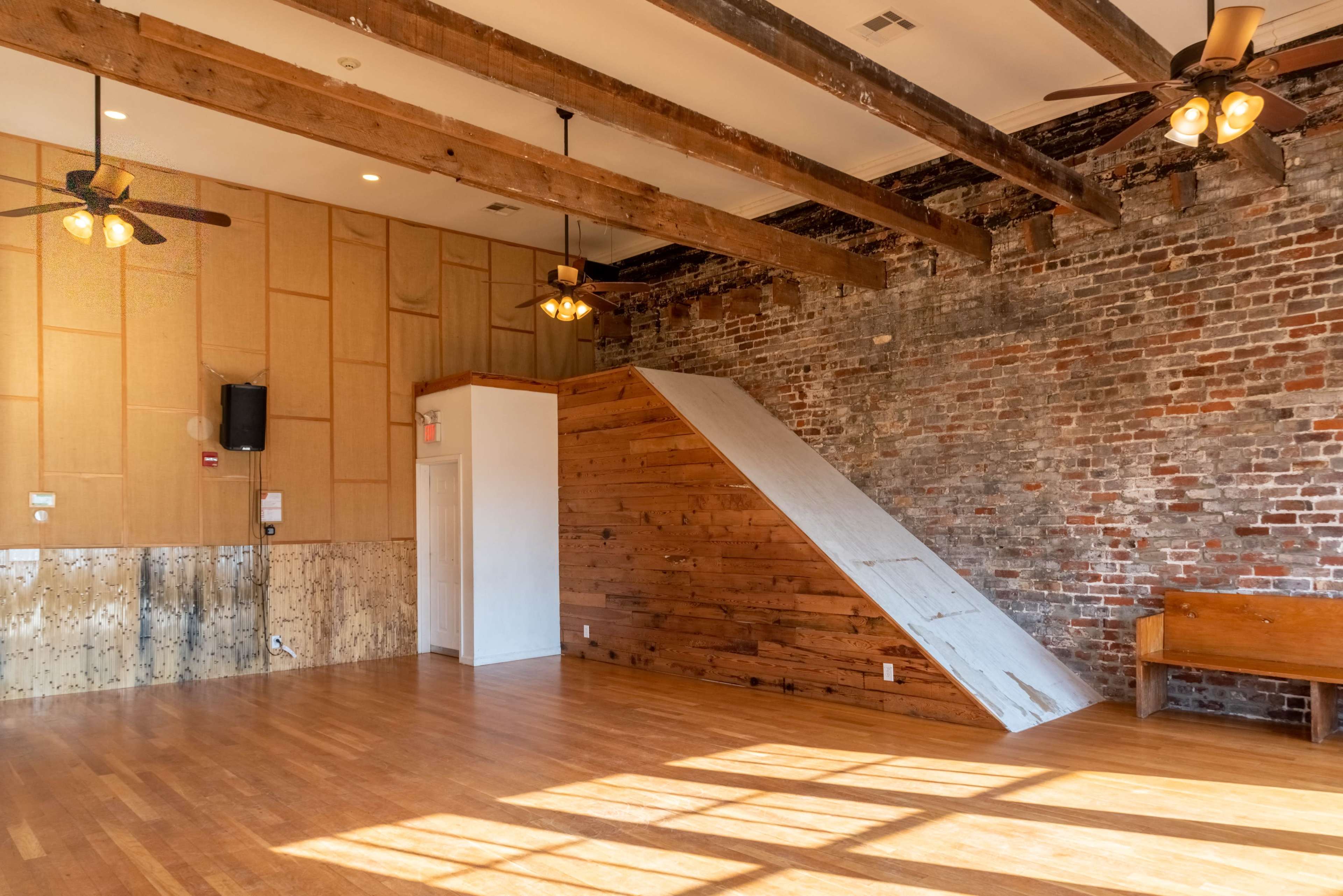 The image shows a spacious interior with exposed brick walls, wooden beams, and a sloped wooden structure leading to a raised area.