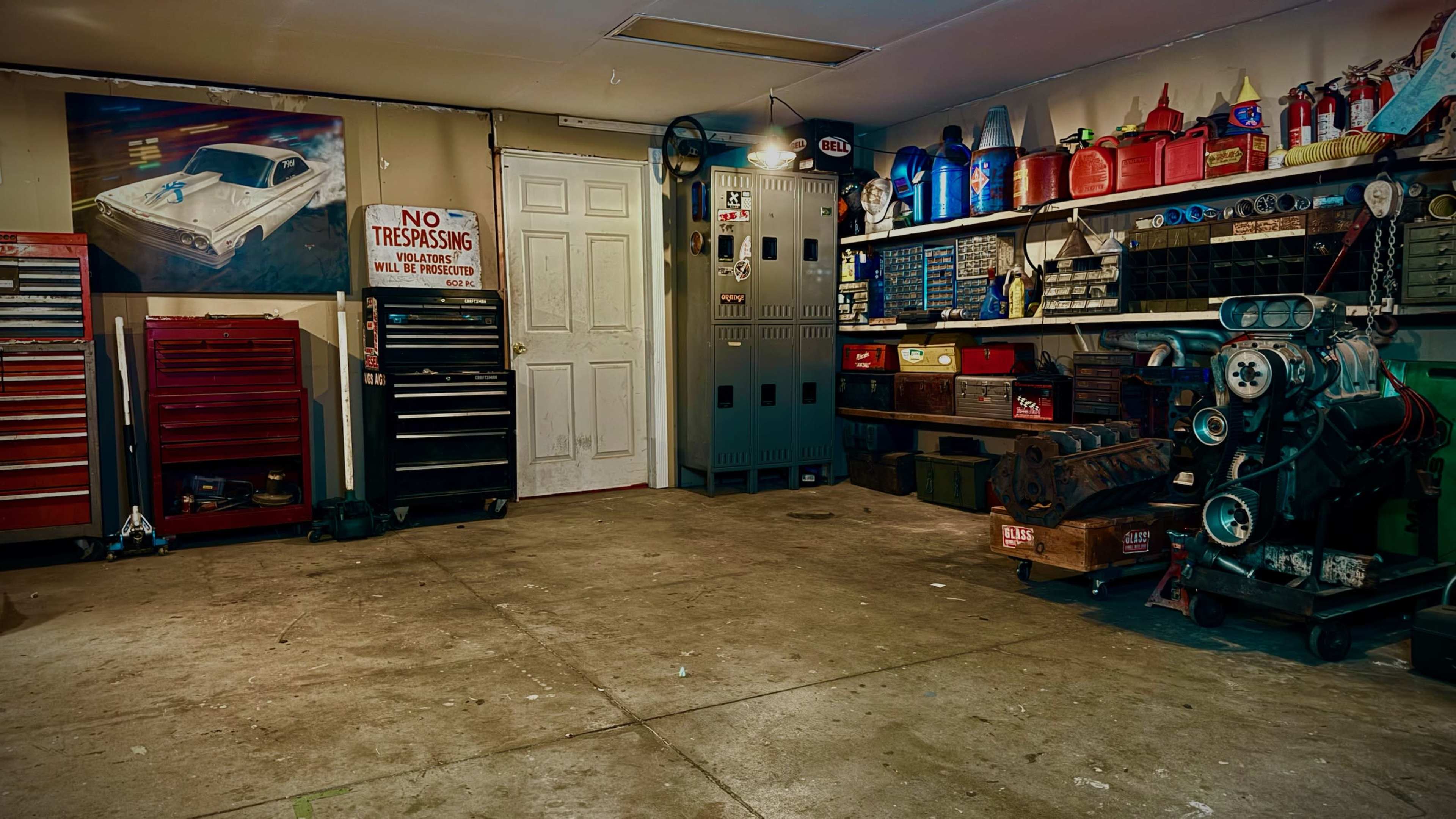 The image shows a garage with tool storage units, a workbench, a vintage car poster on the wall, and various containers and equipment arranged on shelves.