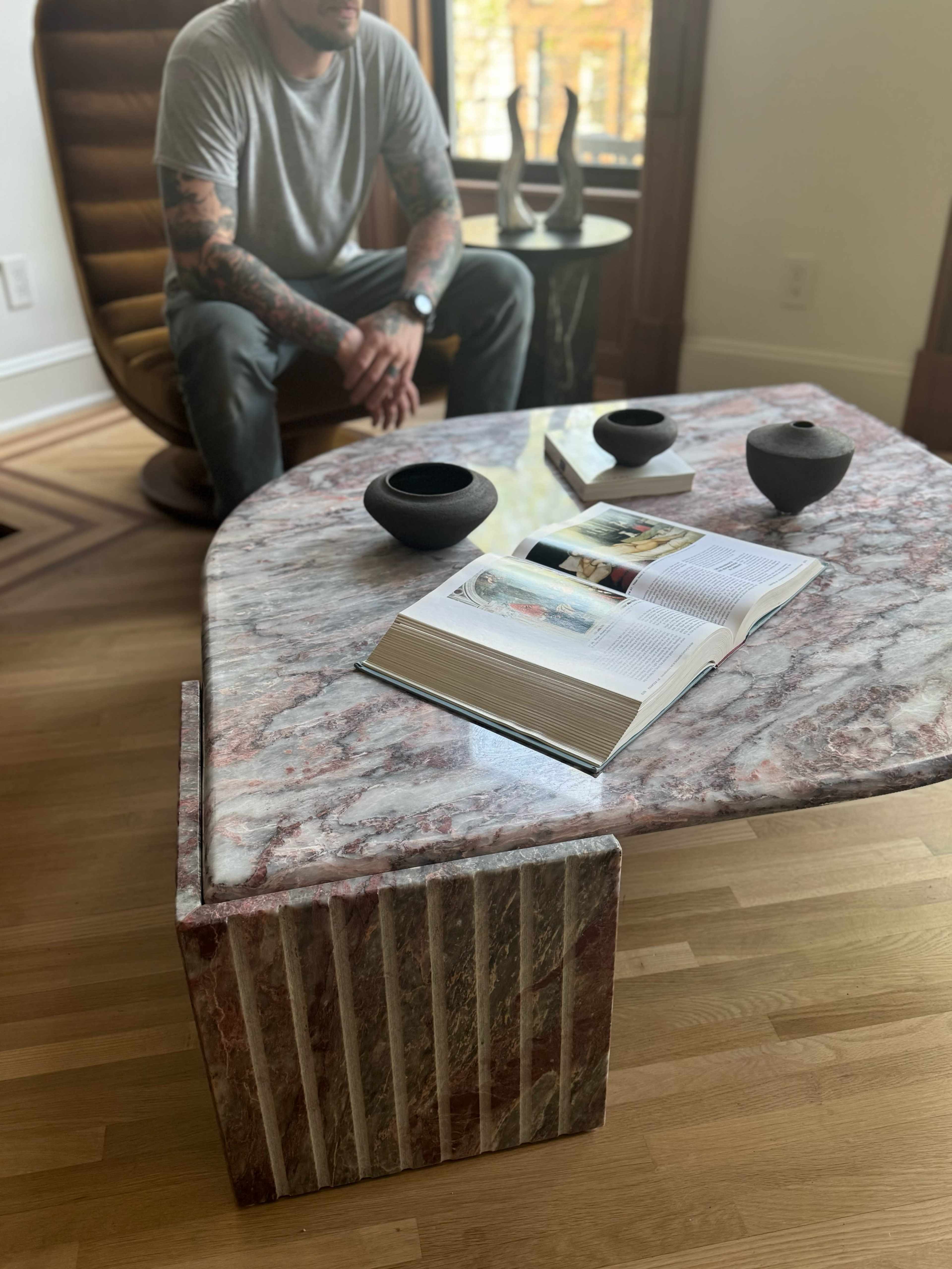 A person with tattoos sits next to a marble coffee table featuring an open book and three black bowls in a modern living space.