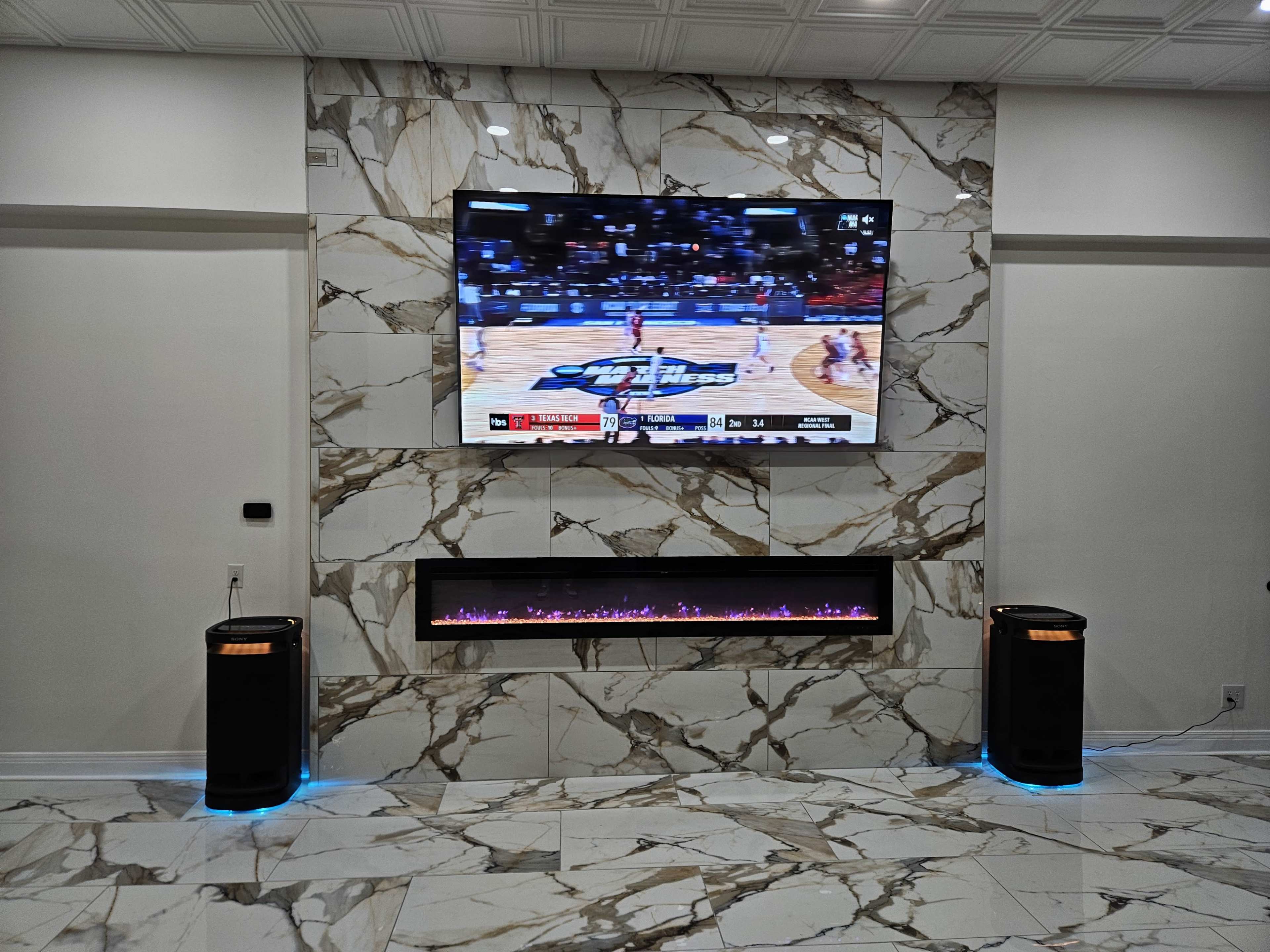 A large flat-screen TV displaying a basketball game is mounted on a marble wall above a modern electric fireplace with blue lighting.
