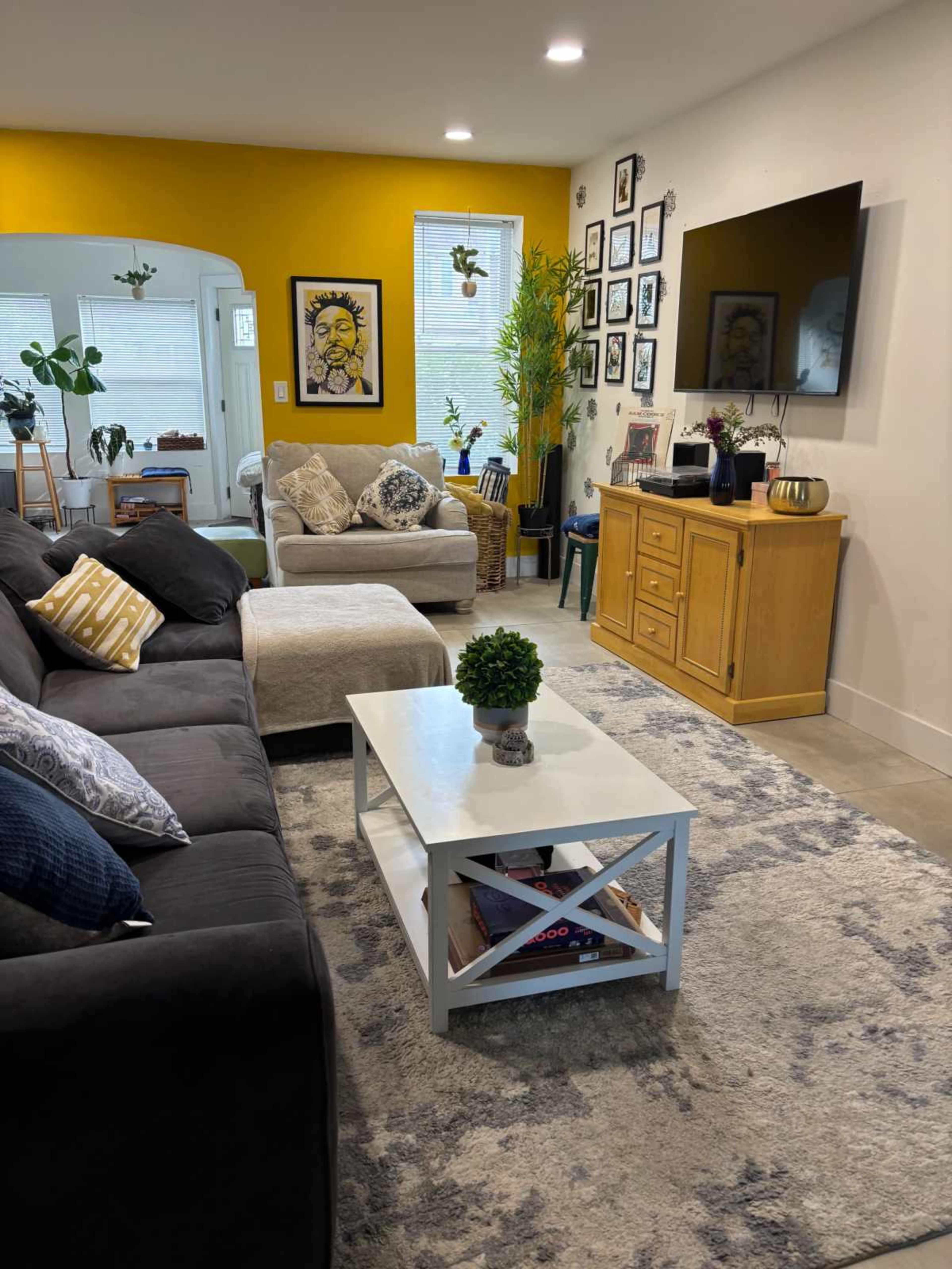 The image shows a cozy living room with a dark sofa, a white coffee table, and a blend of plants and framed artwork on the walls.