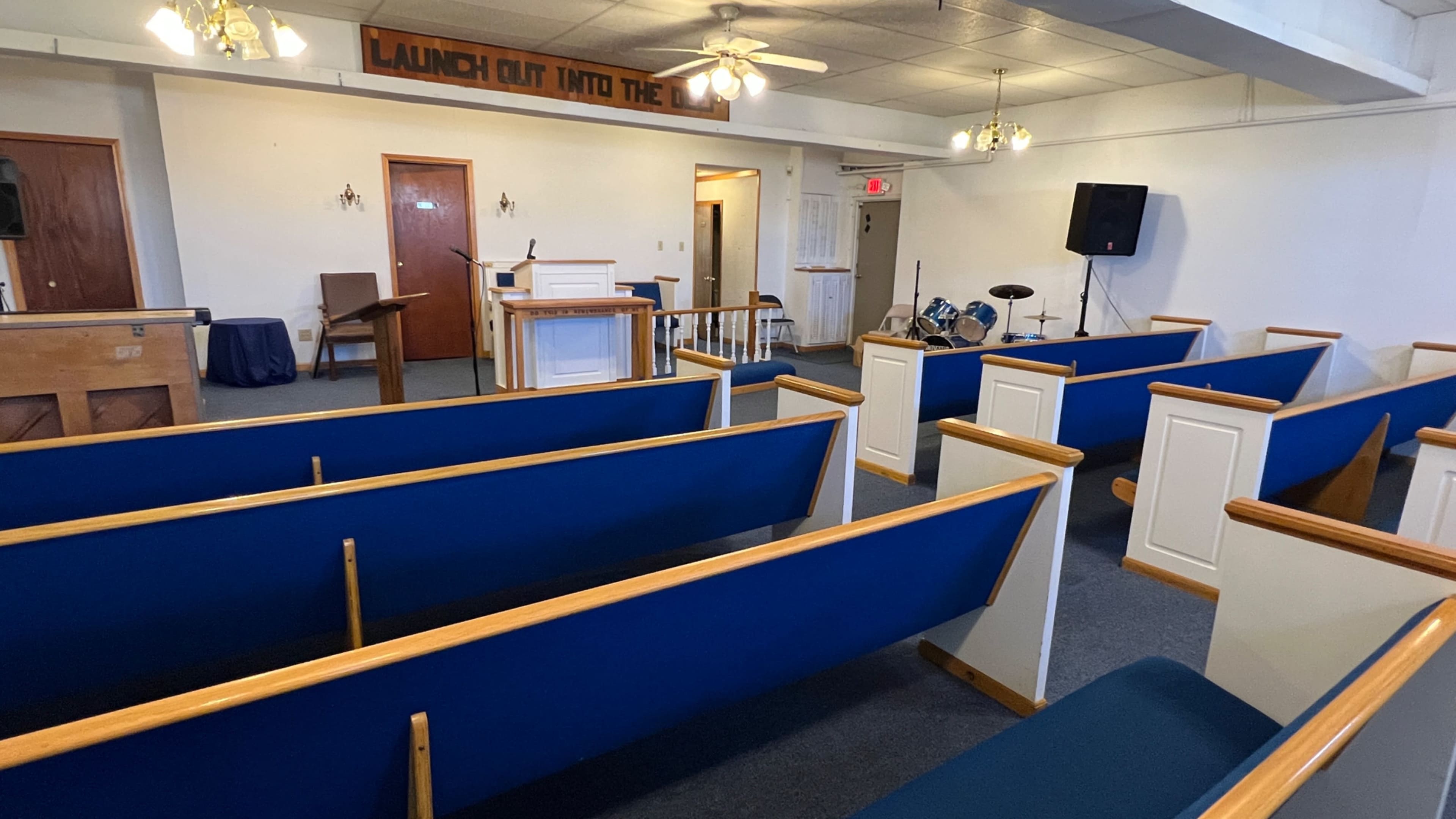 The interior of a church featuring wooden pews with blue cushions, a podium at the front, and a small music setup with drums in the corner.