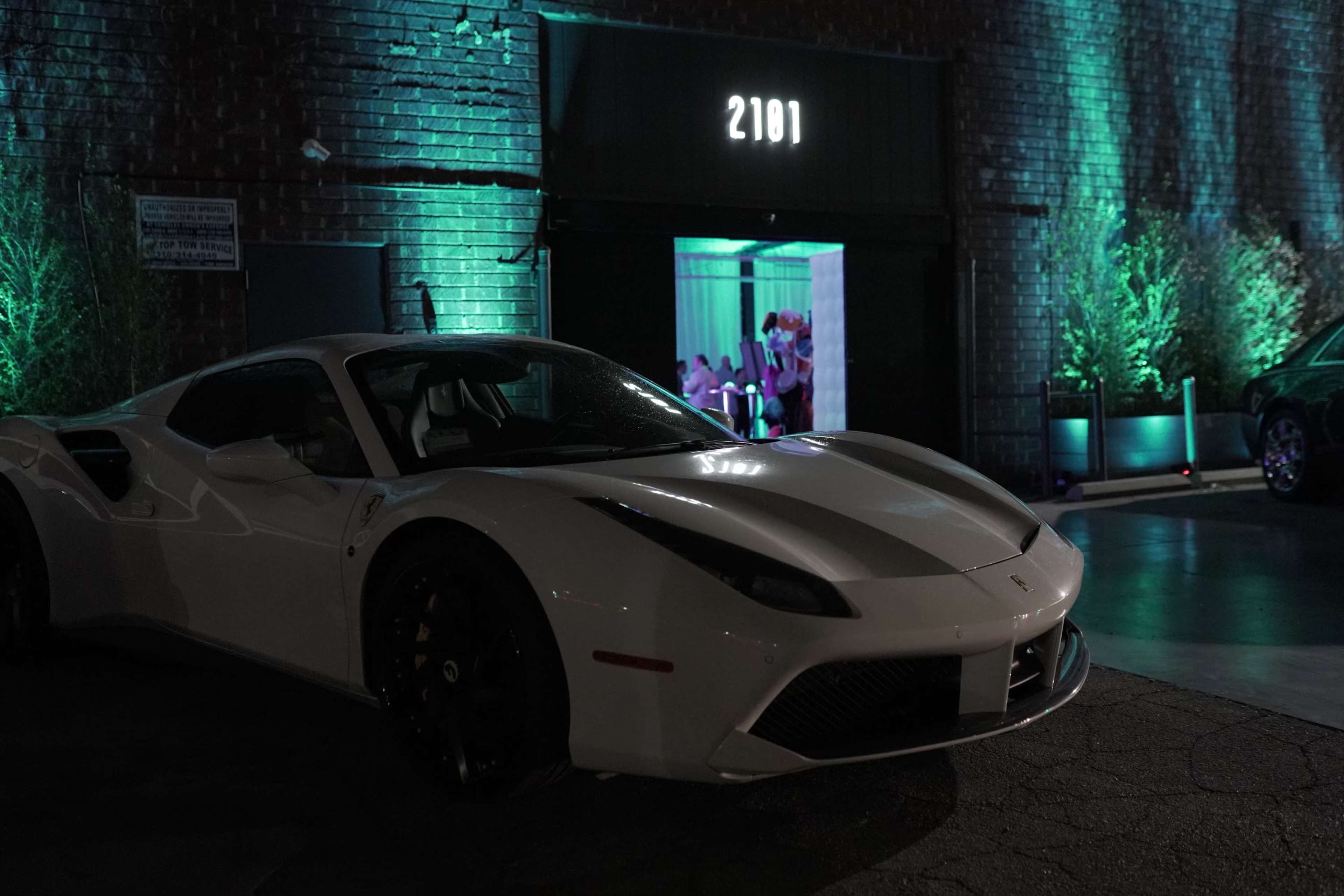A white Ferrari 488 is parked in front of a brick building with teal lighting, showcasing a doorway marked with the number 2101.