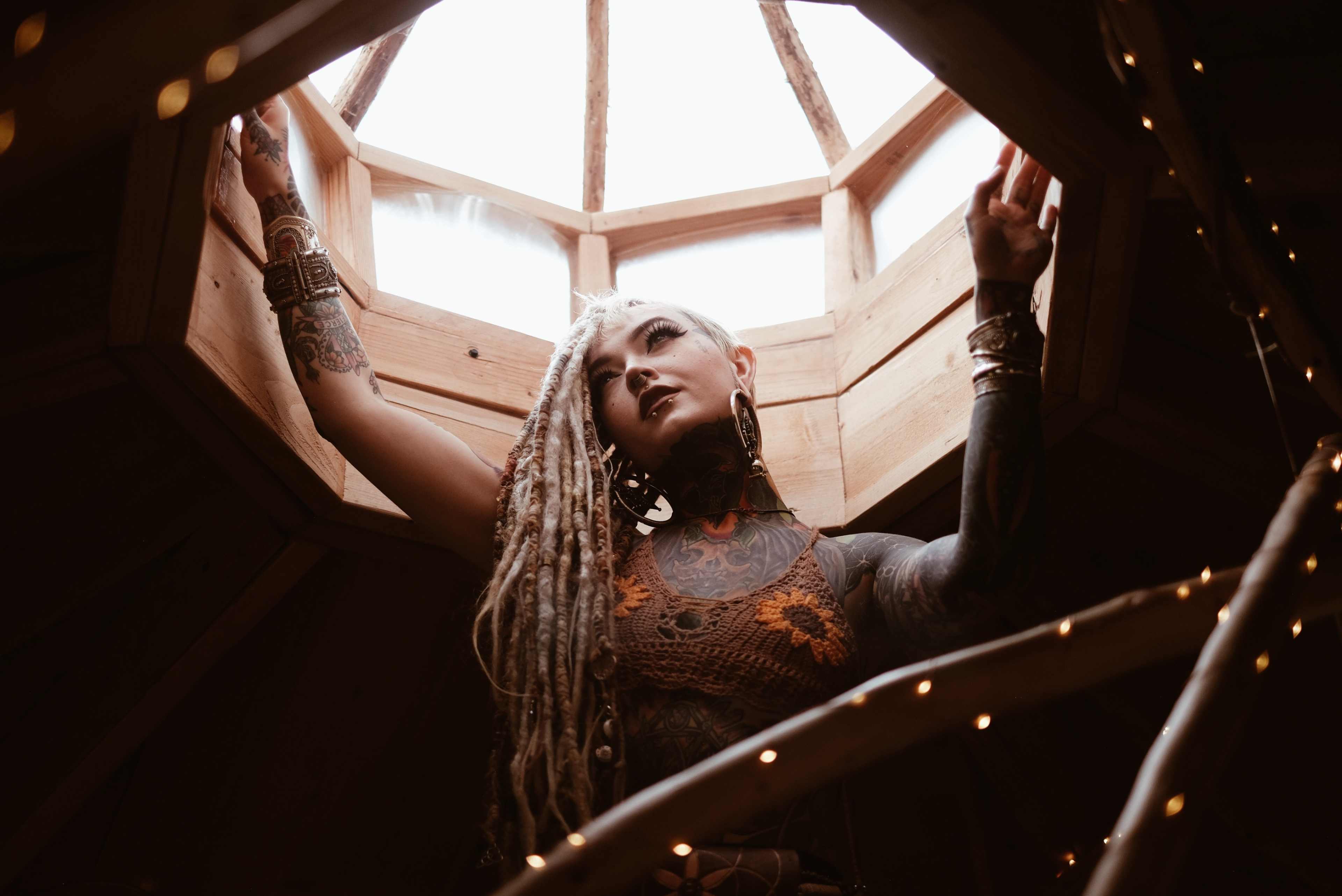 A person with long dreadlocks and tattoos leans against a wooden structure with a skylight above.