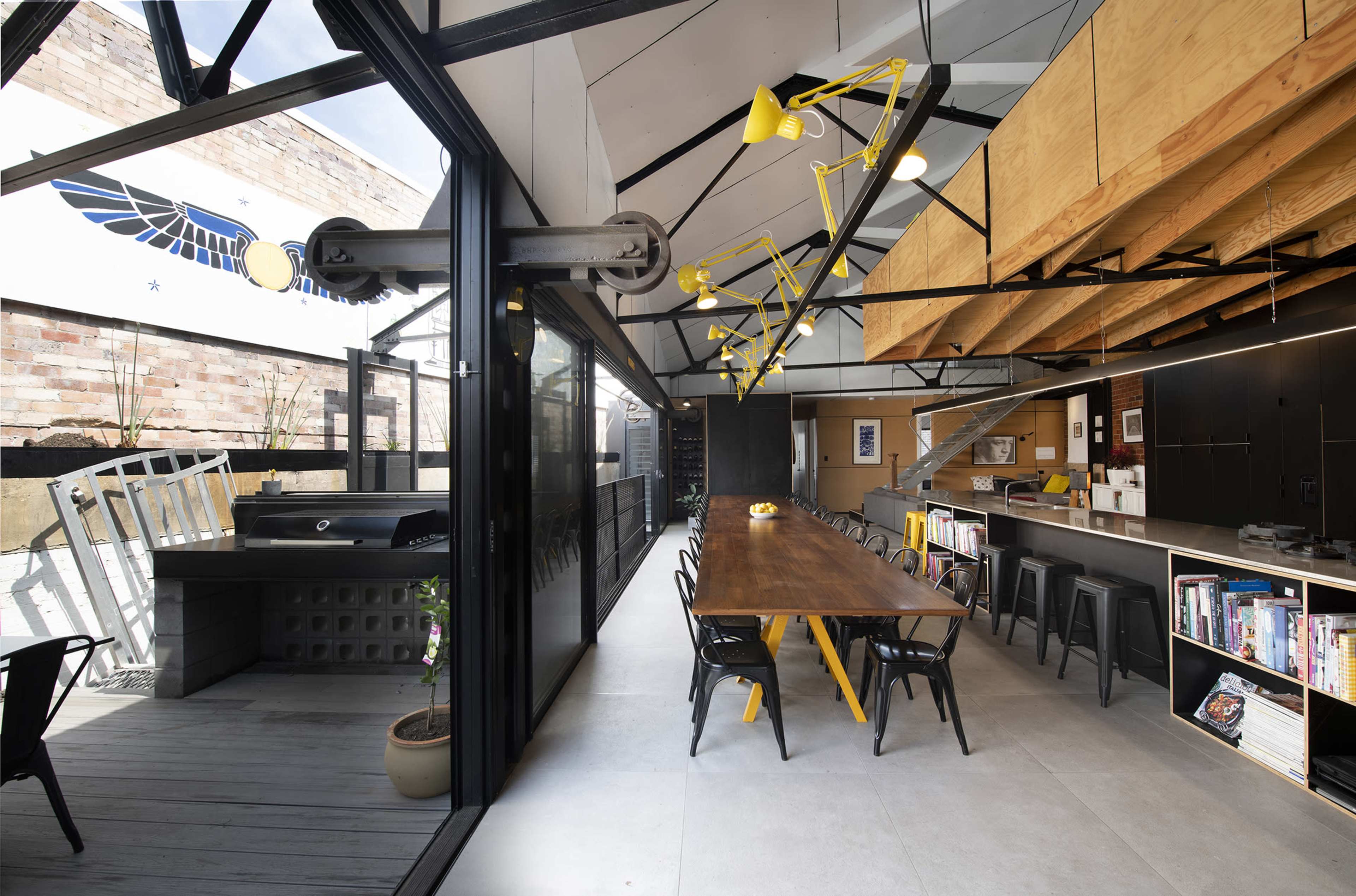 A modern, open-concept interior with a long dining table, black chairs, and large windows, featuring industrial design elements and a striking overhead light fixture.