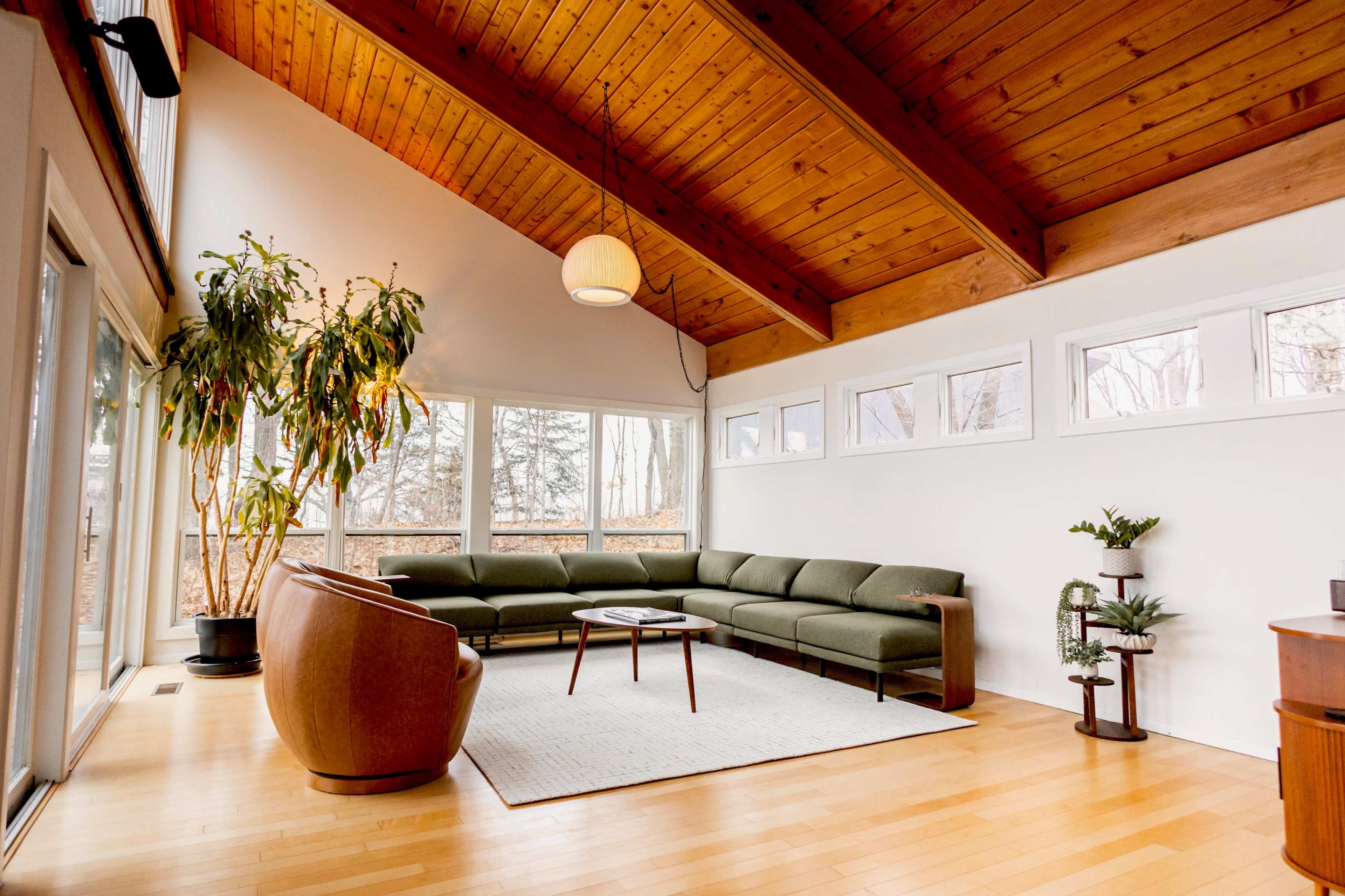 The image features a spacious living room with a brown wooden ceiling, large windows, a green sectional sofa, and a round coffee table on a light area rug.