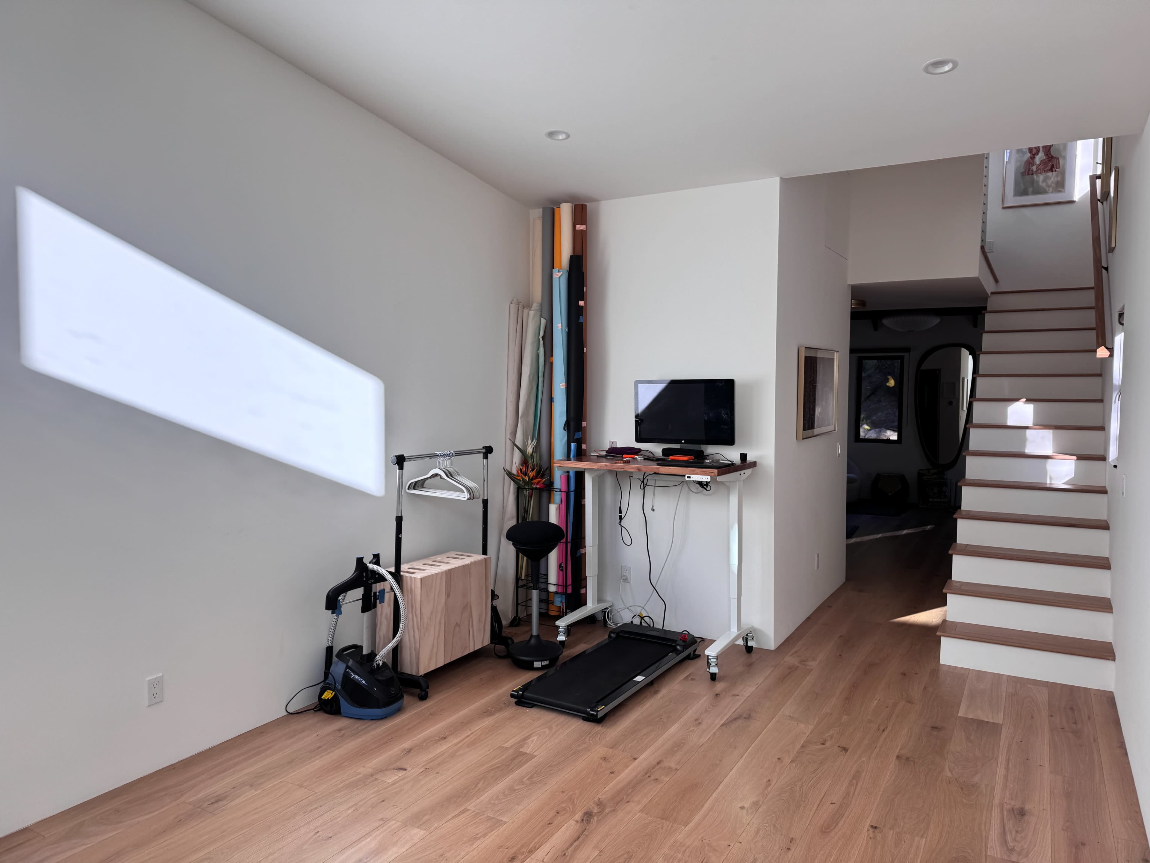 A partially furnished room features a treadmill, a desk with a monitor, and a staircase leading to another level.