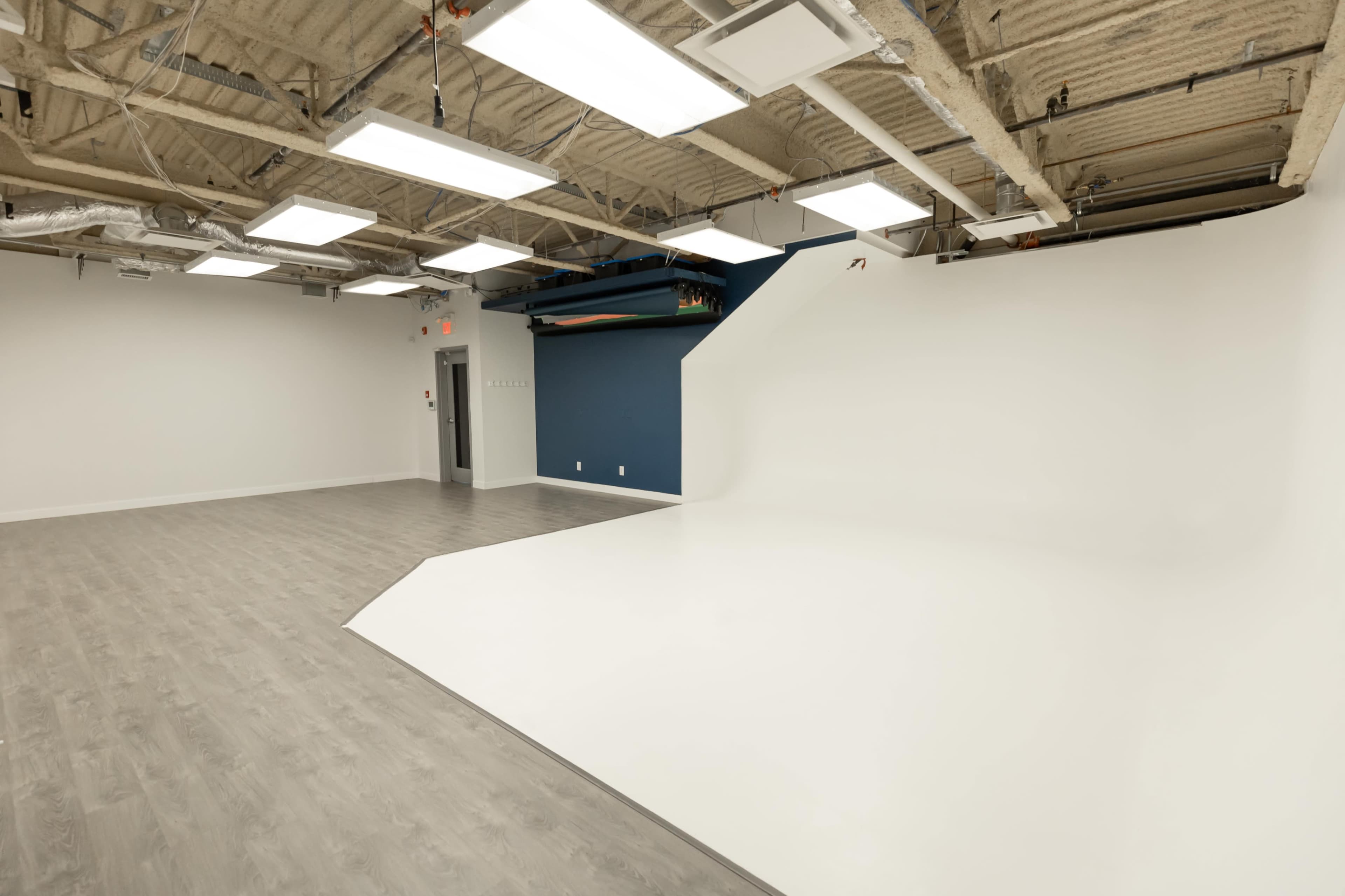 A spacious, empty studio with a white backdrop and gray flooring, featuring overhead lights and exposed ceiling elements.