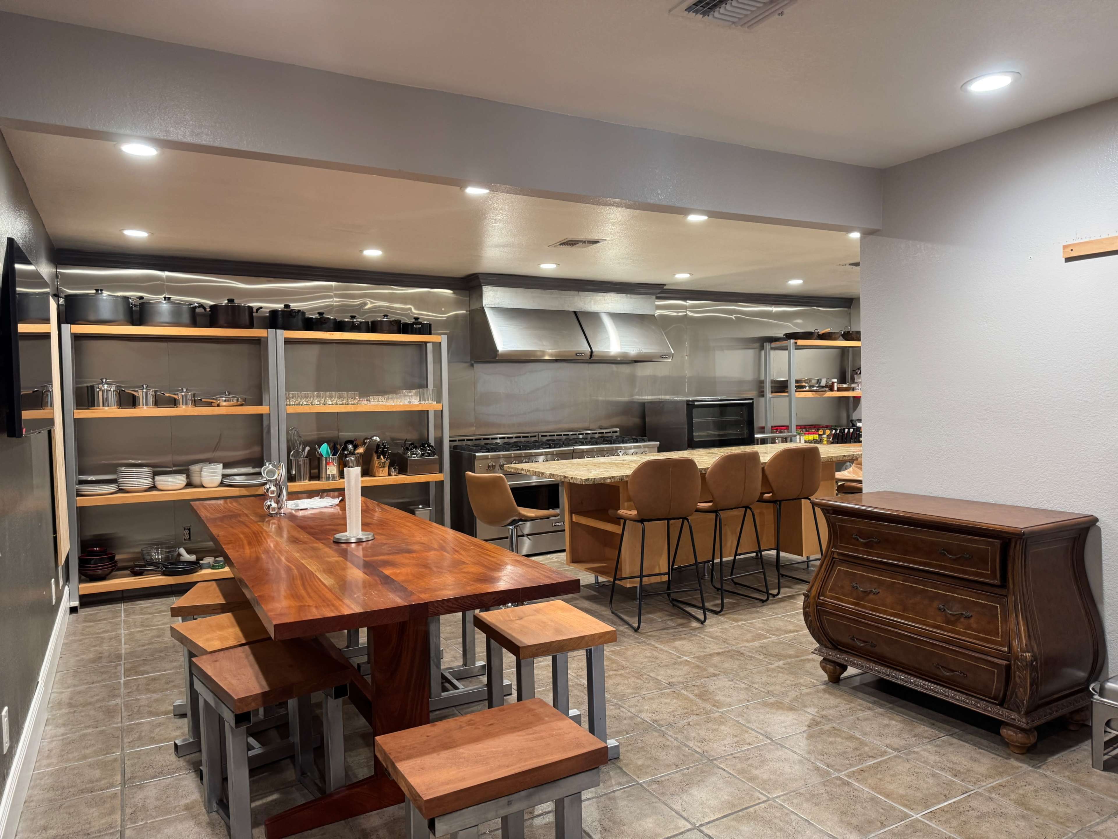 The image shows a spacious kitchen with a large wooden table and benches on one side, bar seating at a counter, and open shelving displaying kitchenware and utensils.