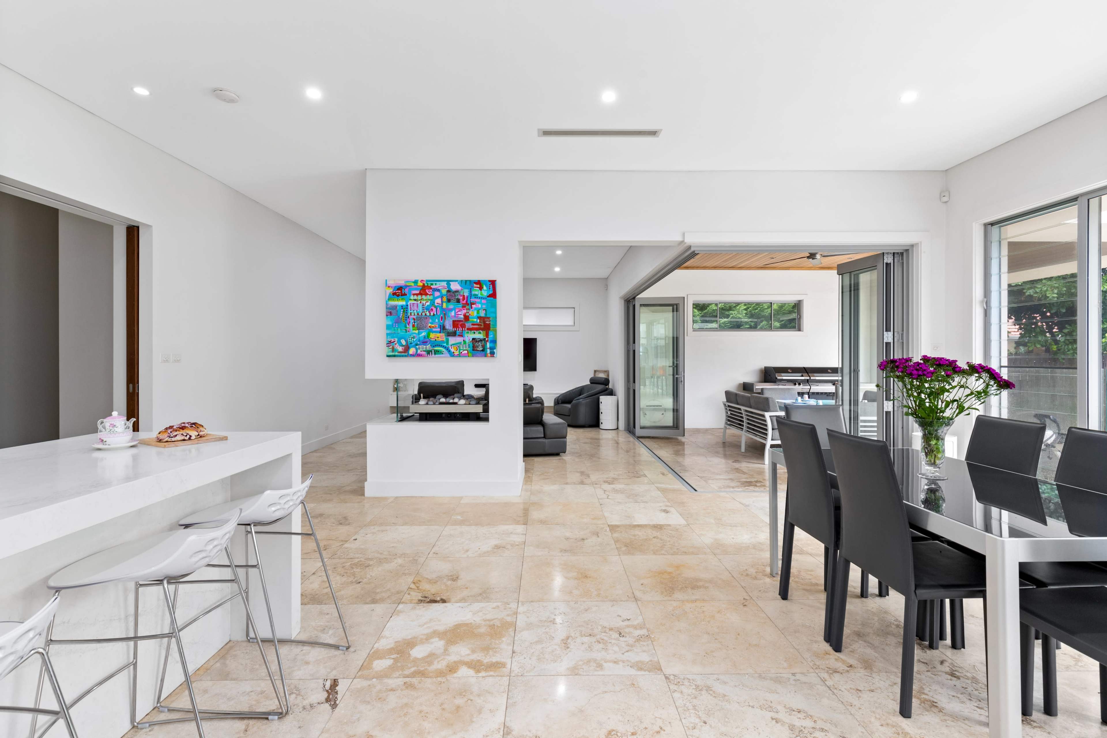 The image shows a modern open-plan living space featuring a dining area, a kitchen counter with stools, and a lounge area with large windows.