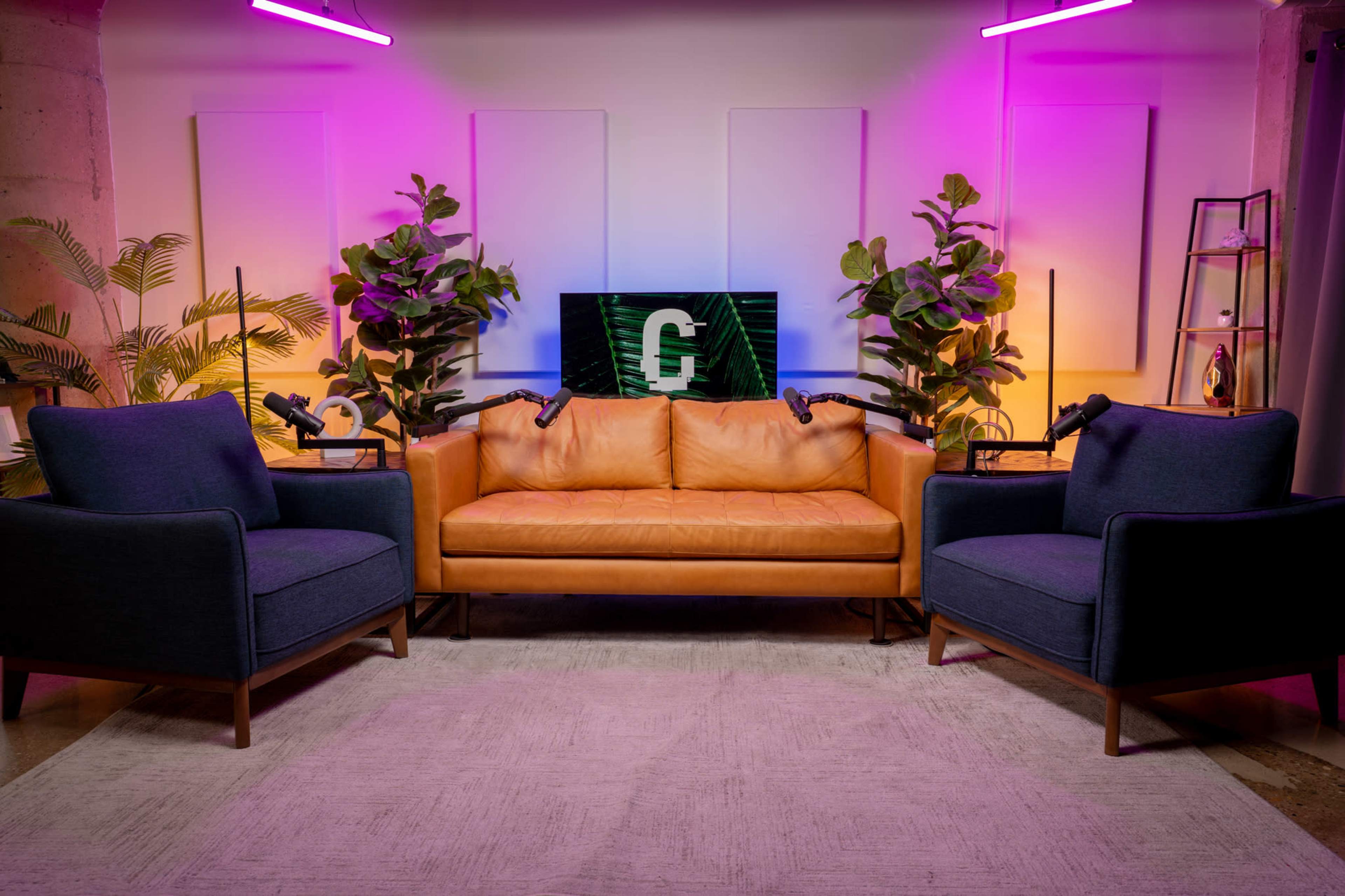 A modern living space features a tan leather sofa flanked by two dark blue armchairs, with potted plants and a television displaying a logo in the background under colorful lighting.