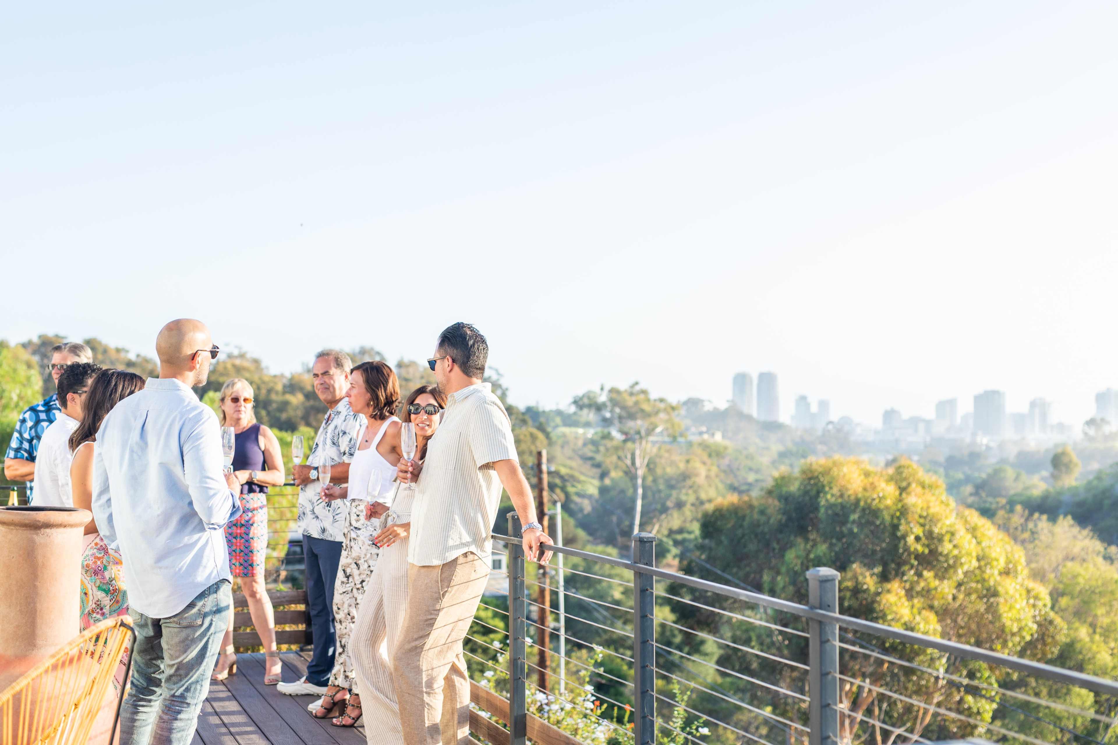 A group of people gathers on a deck overlooking a city skyline surrounded by trees during a daytime event.