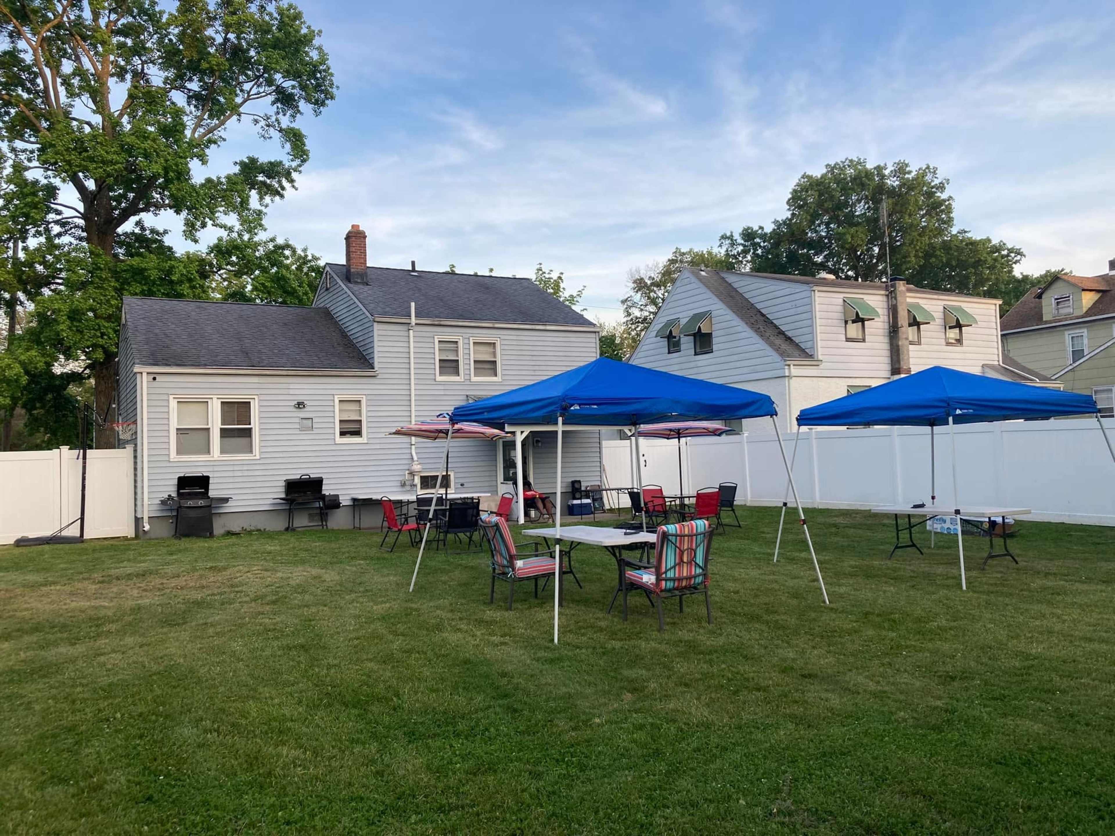 A grassy backyard features two blue canopies, several tables and chairs, and two houses with gray siding in the background.