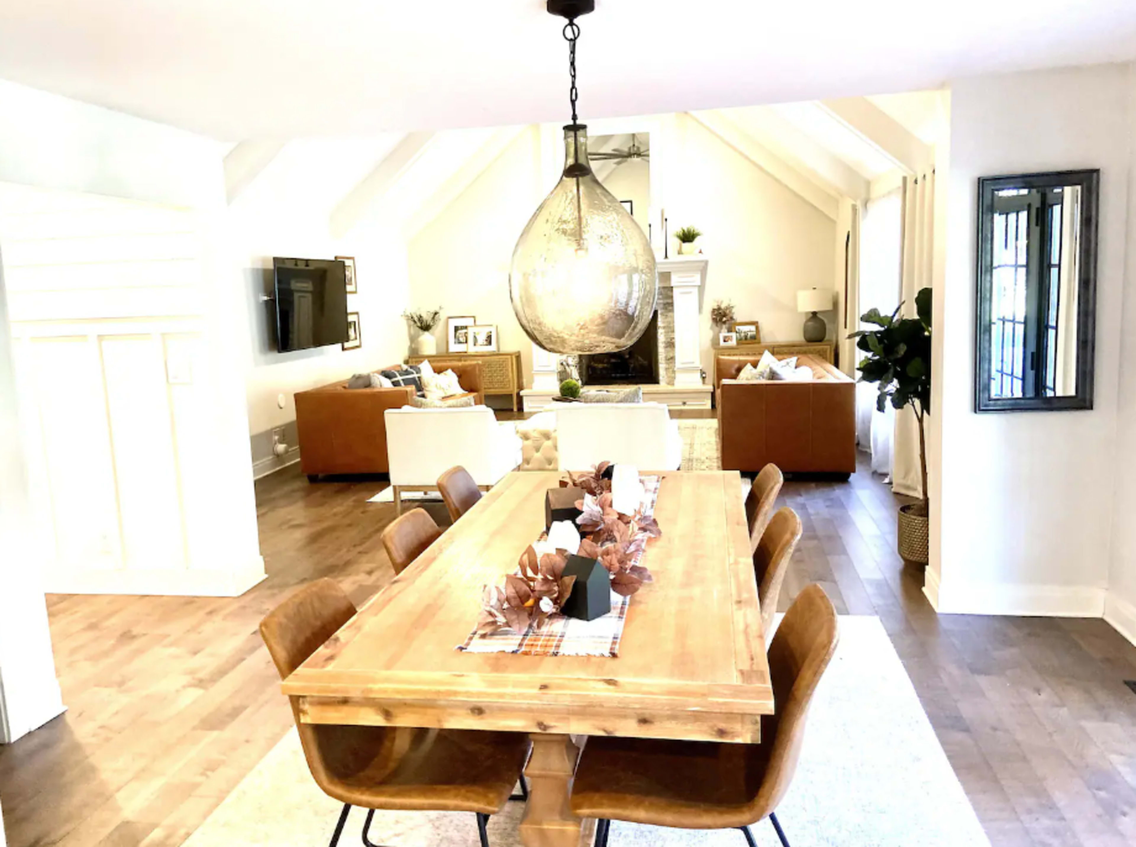 The image shows a spacious dining area with a long wooden table surrounded by brown chairs, leading into a cozy living room furnished with light-colored sofas and a large wall-mounted TV.