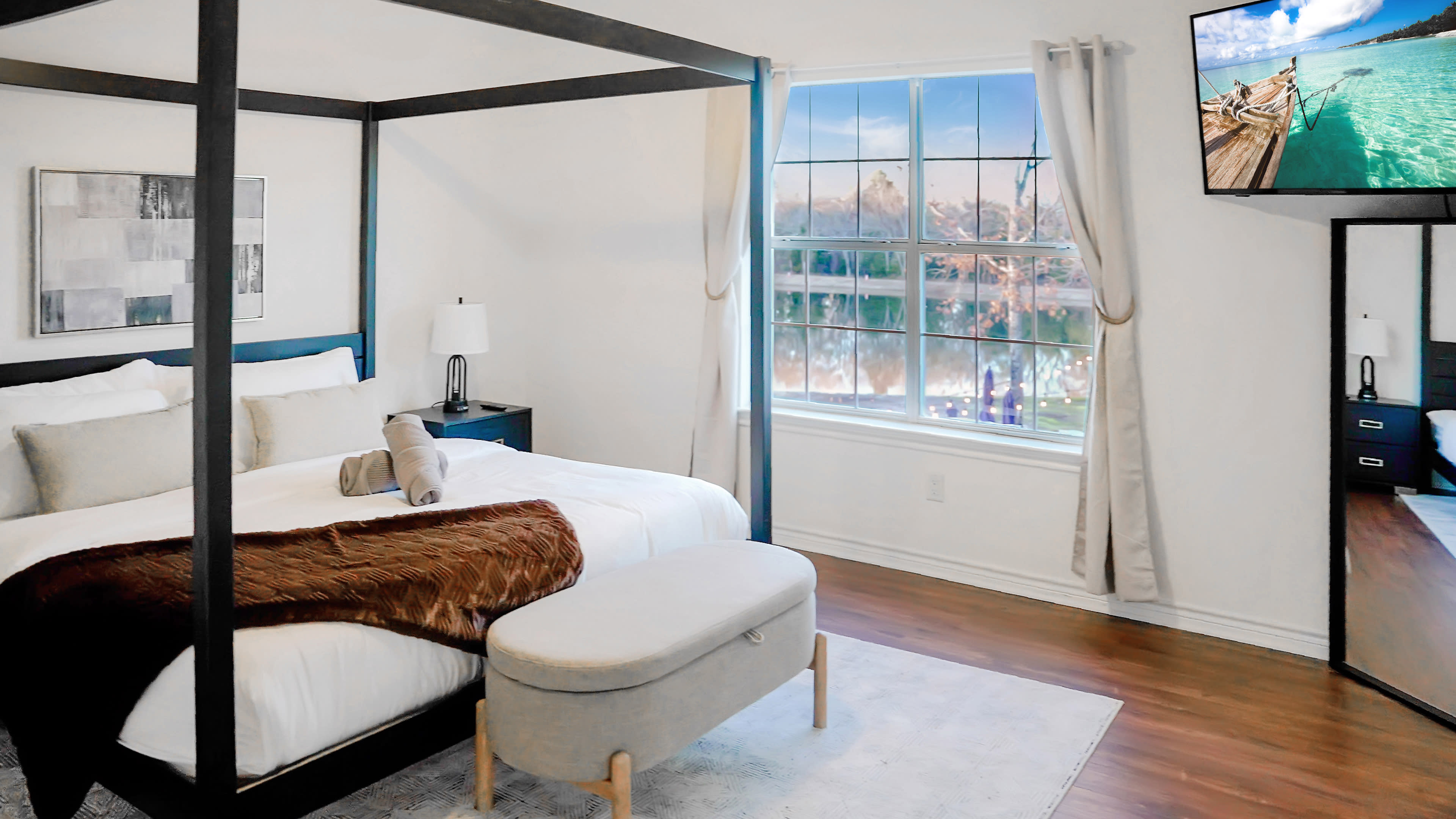 The image shows a modern bedroom featuring a four-poster bed, a bench at the foot, a large window with a view, and a television mounted on the wall.