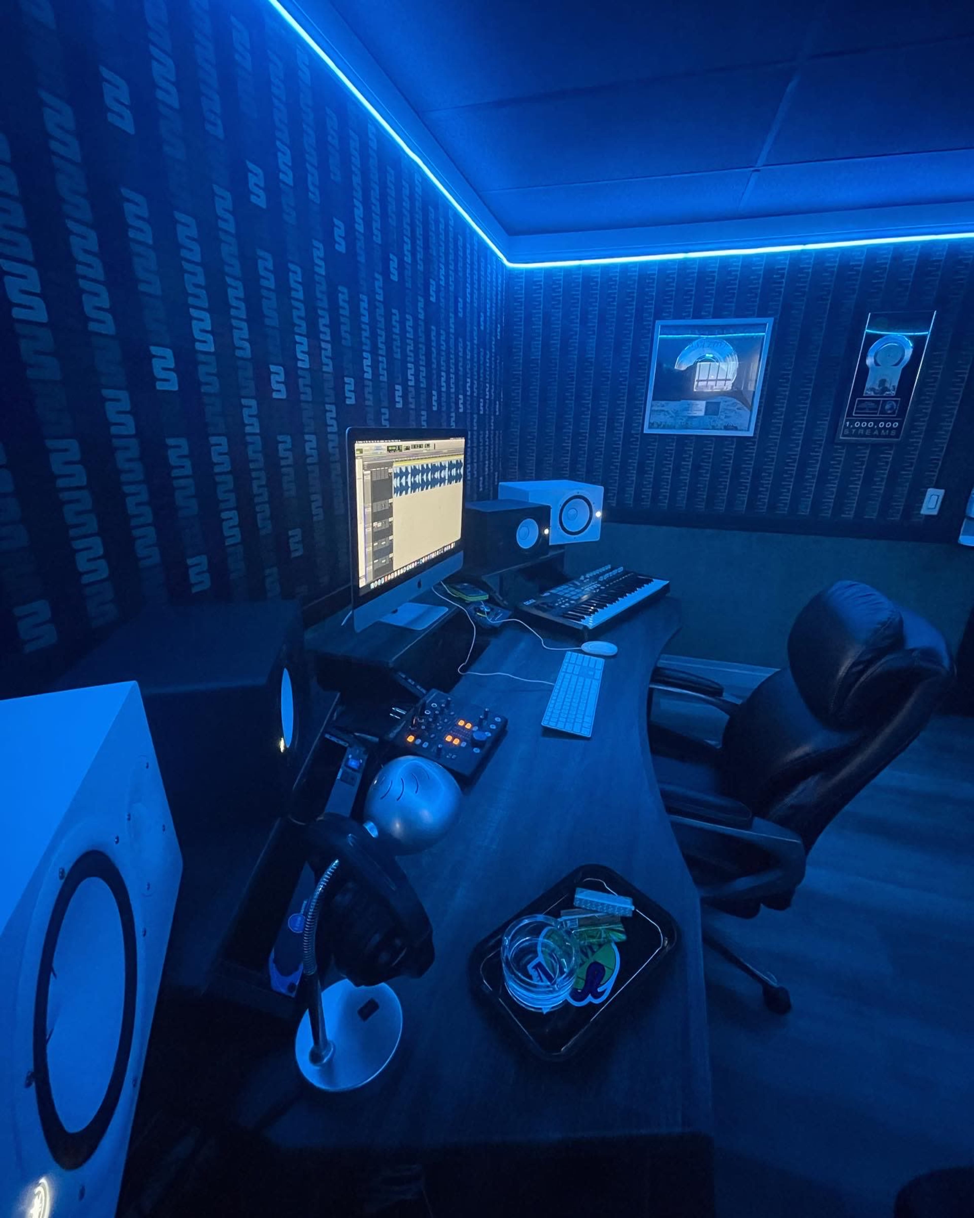 A modern music studio features a desk with a computer, audio interface, synthesizer, and speakers, all illuminated by blue lighting.